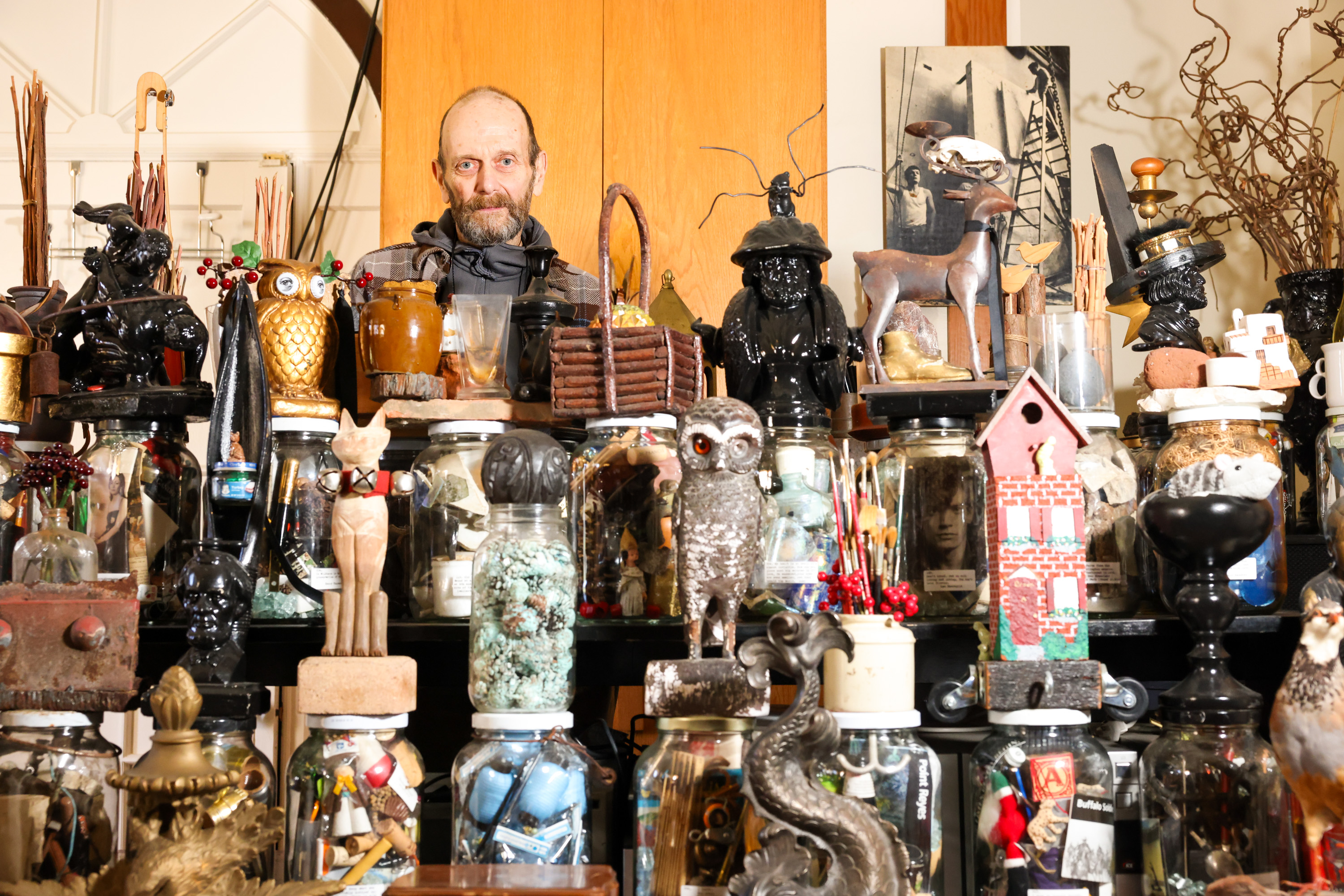 A man stands amidst a cluttered collection of eclectic items, including jars, sculptures, and figurines, like owls and a deer, creating a curious display.