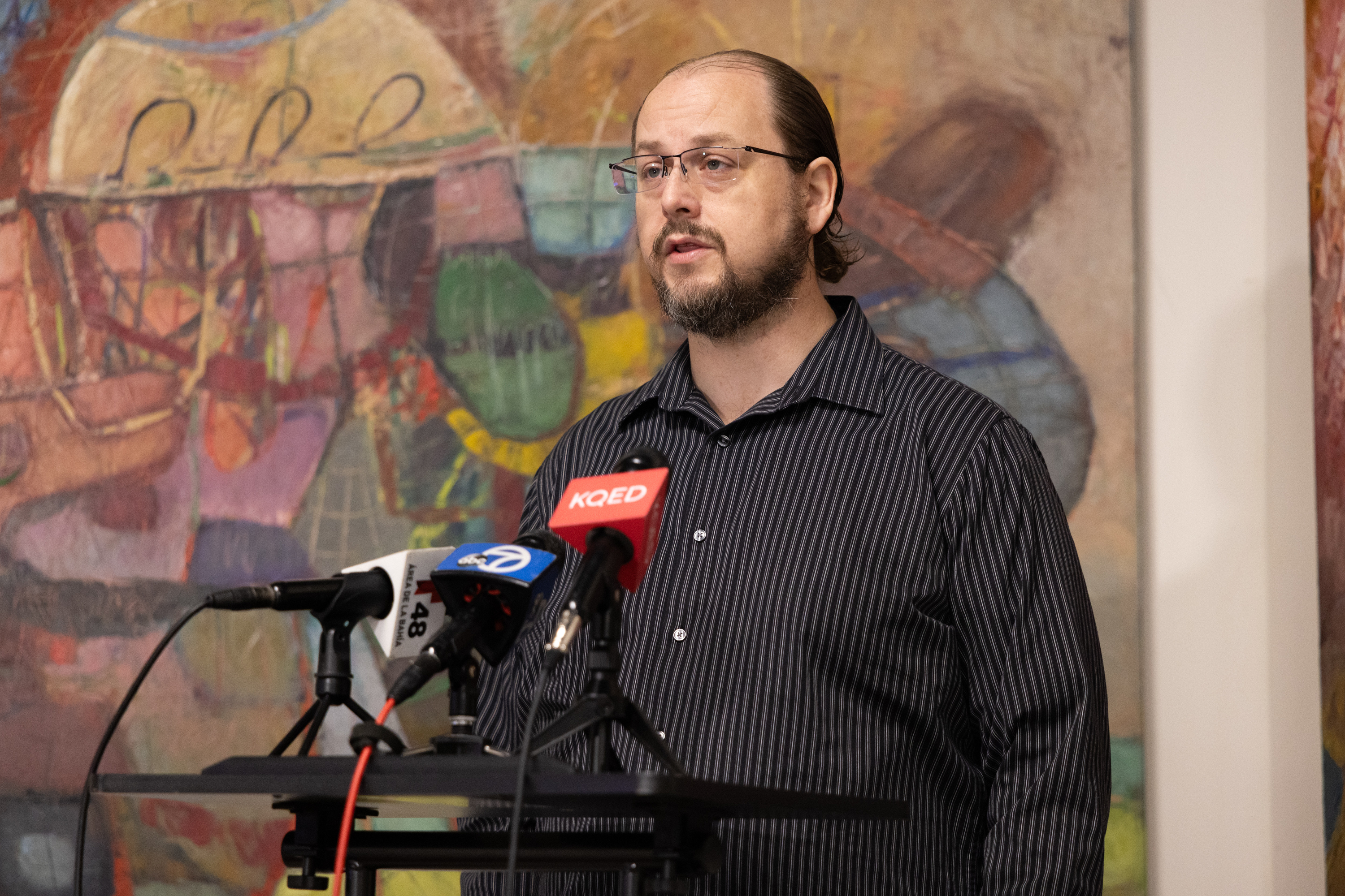 A bearded man in glasses and a striped shirt speaks at a podium with multiple microphones, against a colorful abstract painting background.