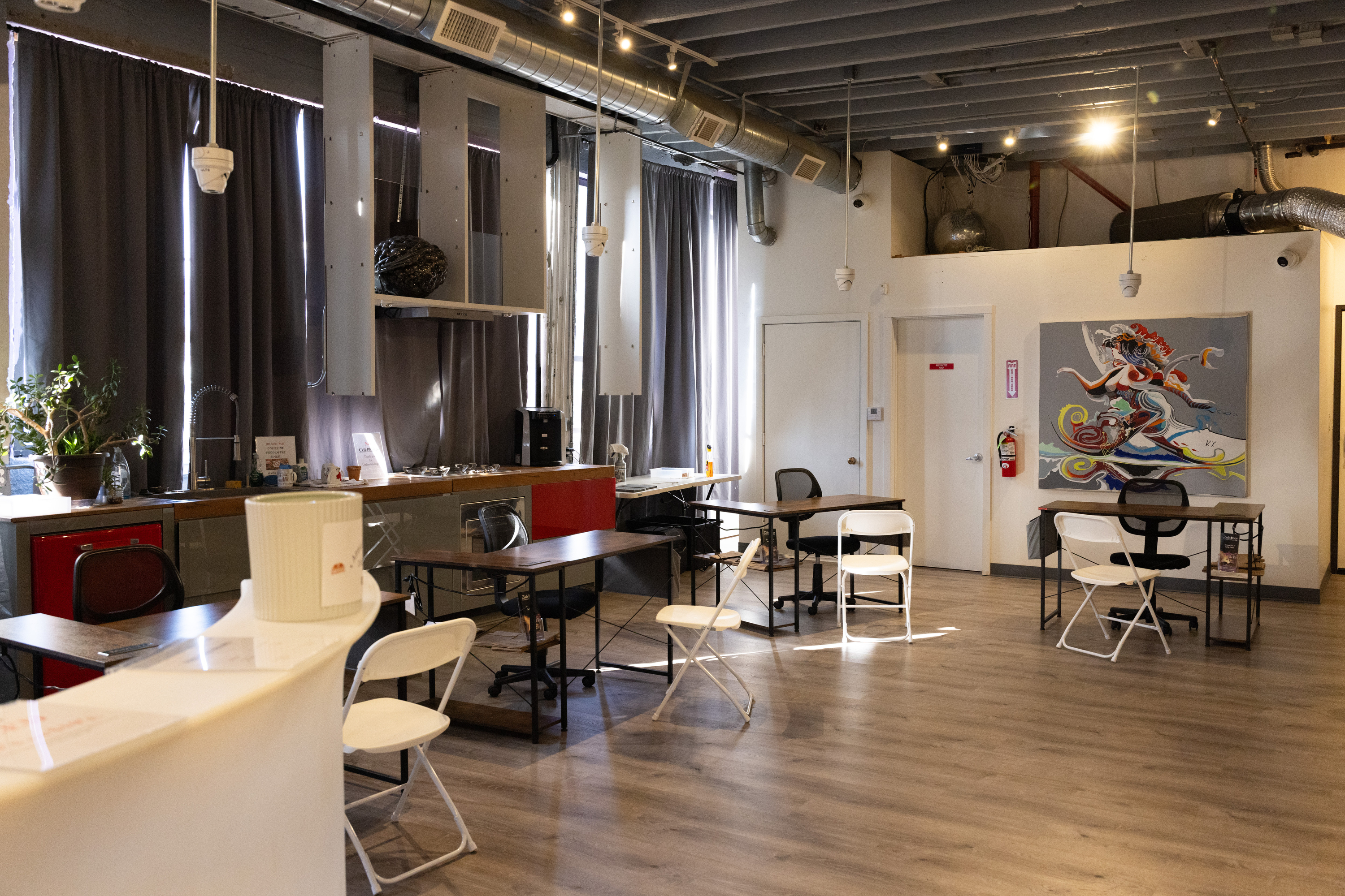 The image shows a modern indoor space with wooden floors, tables, and chairs. There are plants, gray curtains, a colorful painting on the wall, and exposed ductwork.