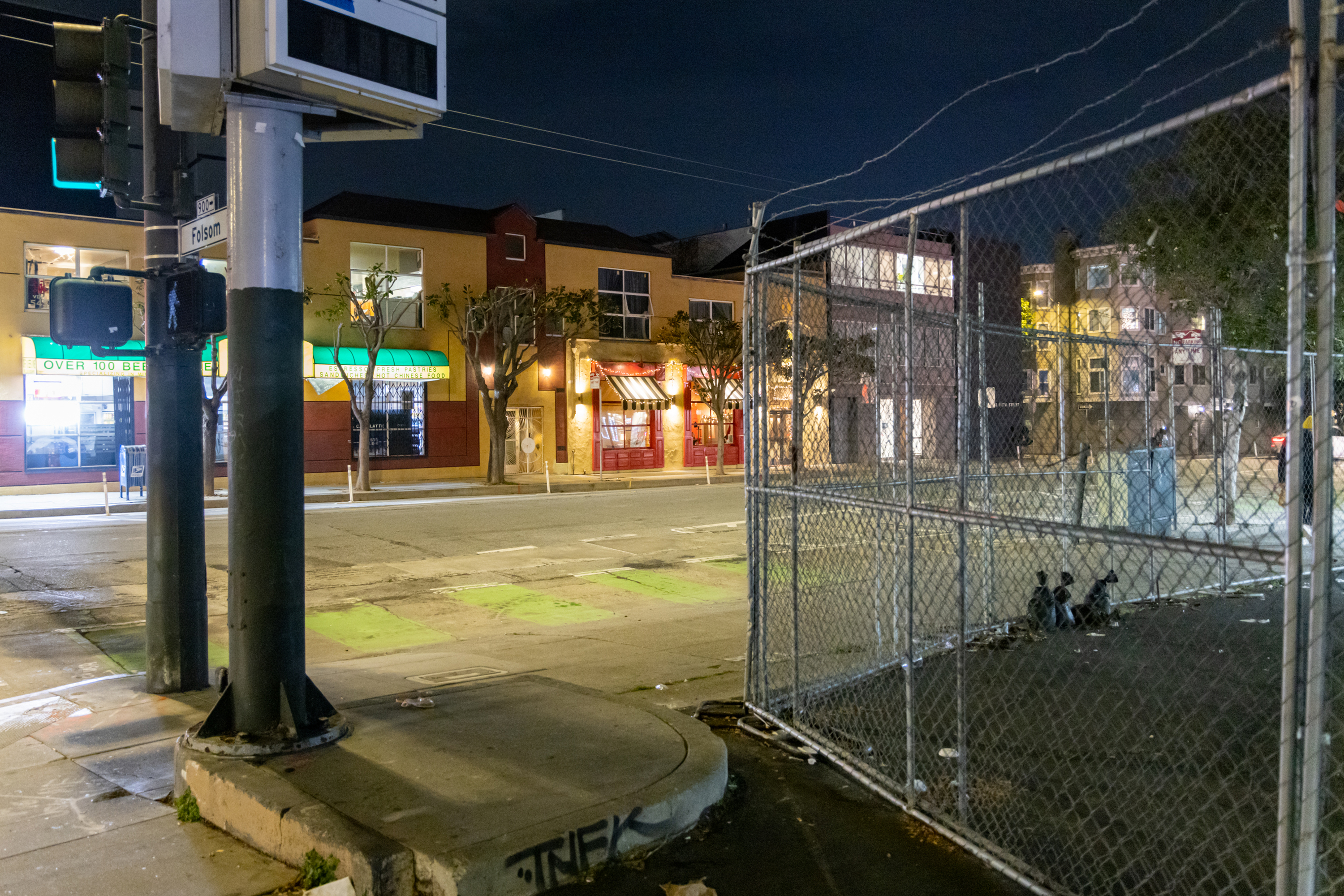 The image shows a city street at night with colorful buildings, including a shop with a green awning. A fenced area is on the right, and streetlights illuminate the scene.