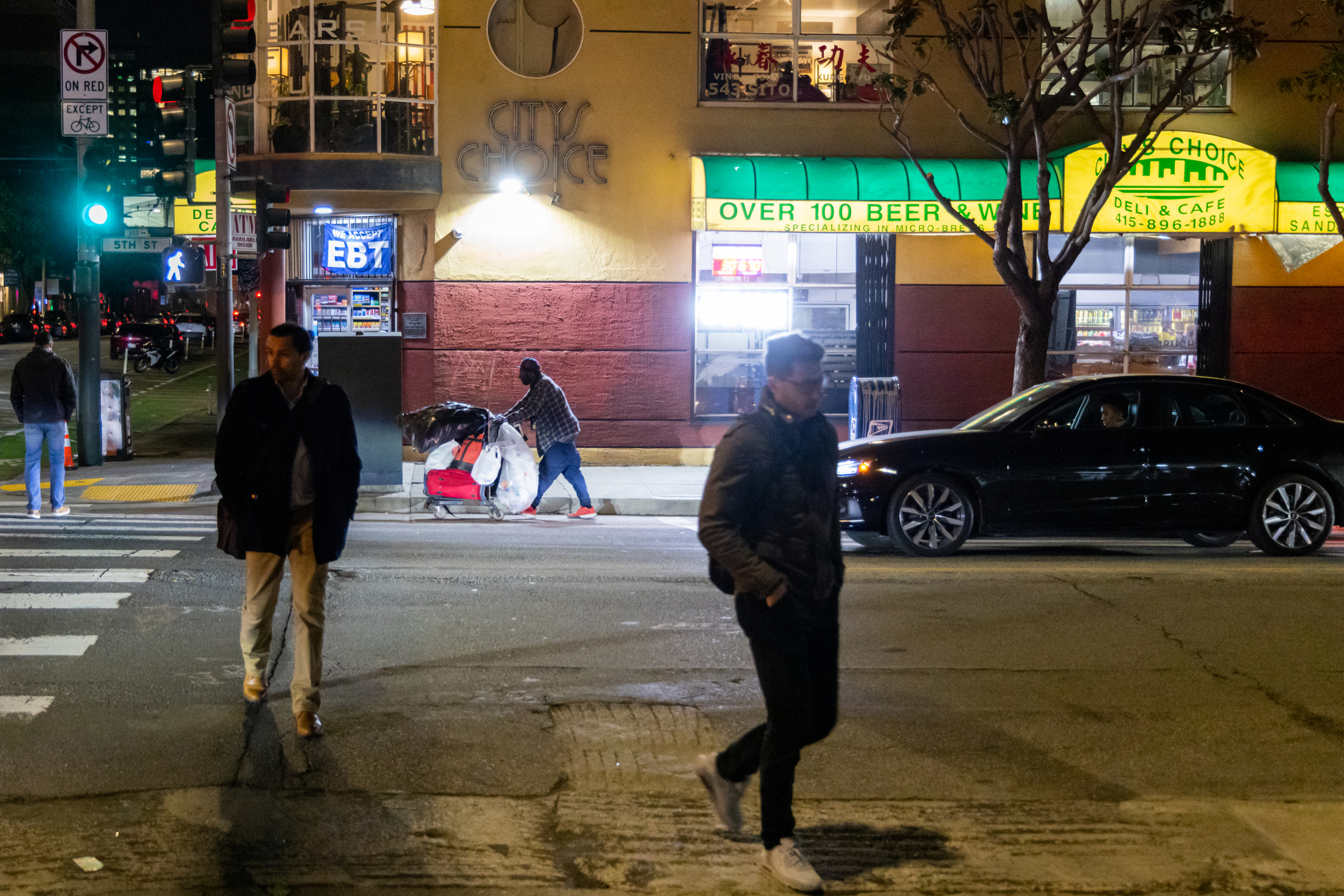 A nighttime street scene shows people walking, a person pushing a cart with bags, and a black car driving past a store with a bright yellow-green awning.