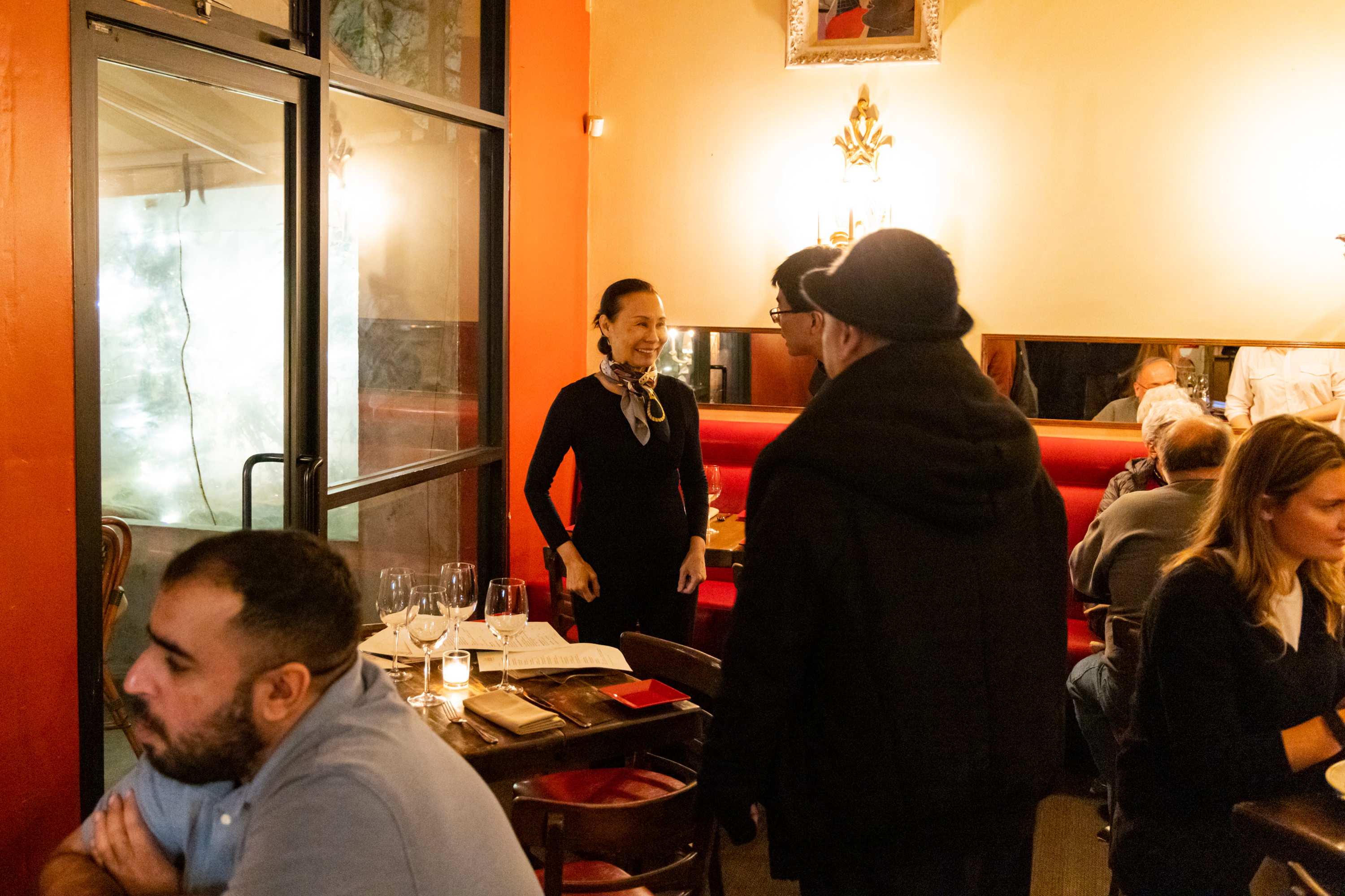 A warmly-lit restaurant shows people dining and conversing. A woman stands smiling, while others sit at tables with wine glasses and candles.