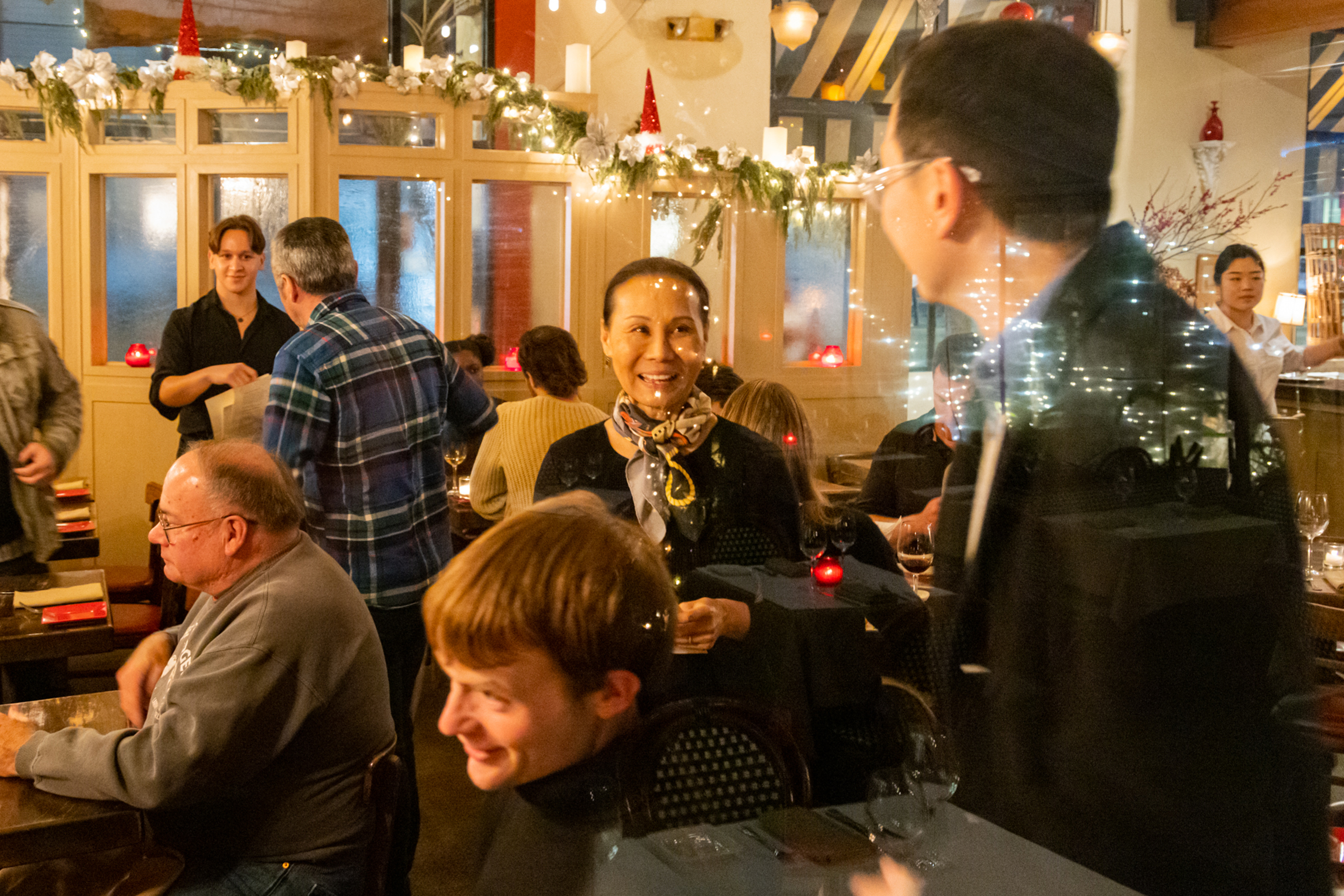 People are enjoying a festive dinner in a decorated restaurant. A woman in a scarf smiles while talking to a man. Holiday lights and greenery adorn the space.