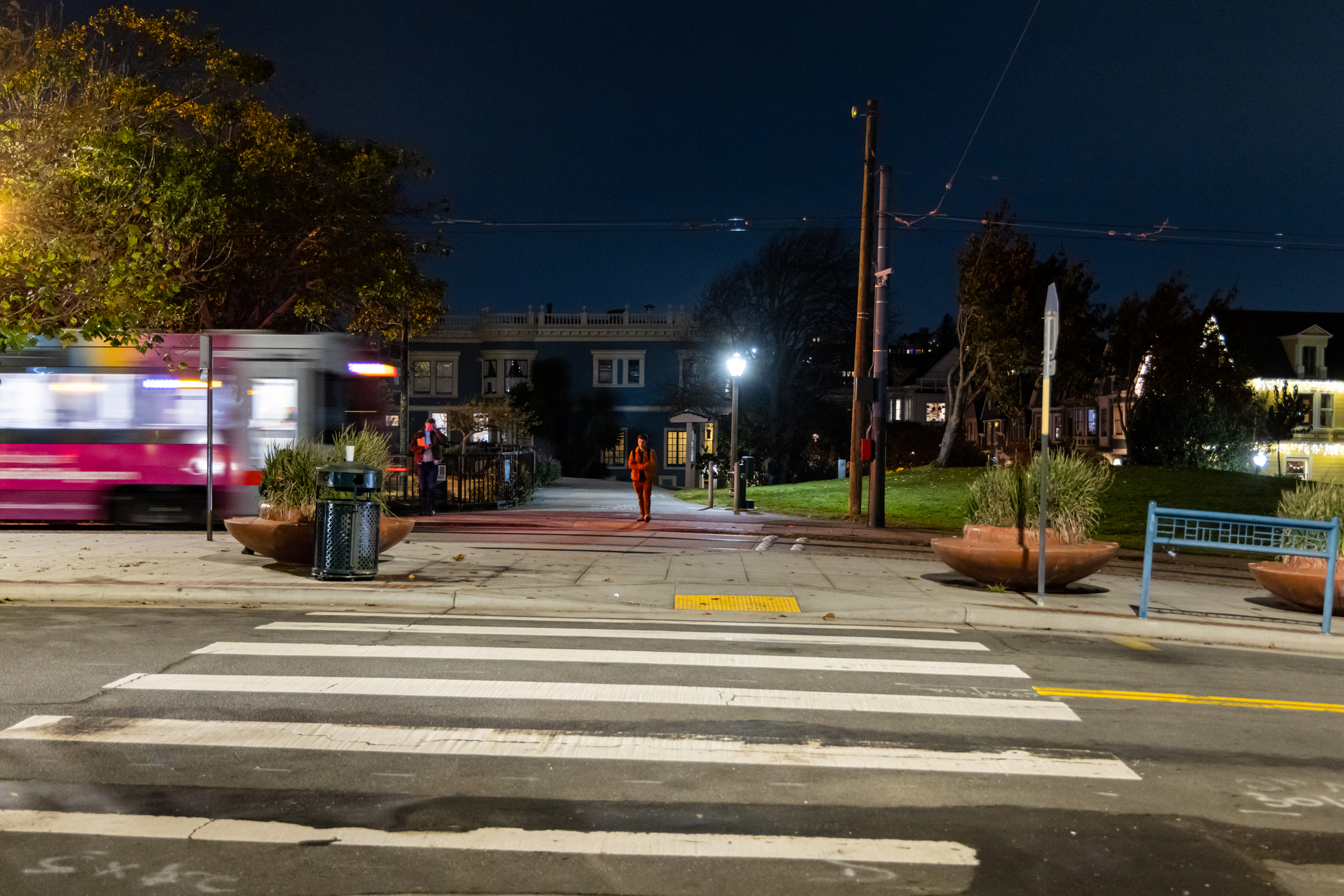 A blurry pink and white tram passes on a street at night. A person walks on the sidewalk under streetlights, with trees and houses in the background.