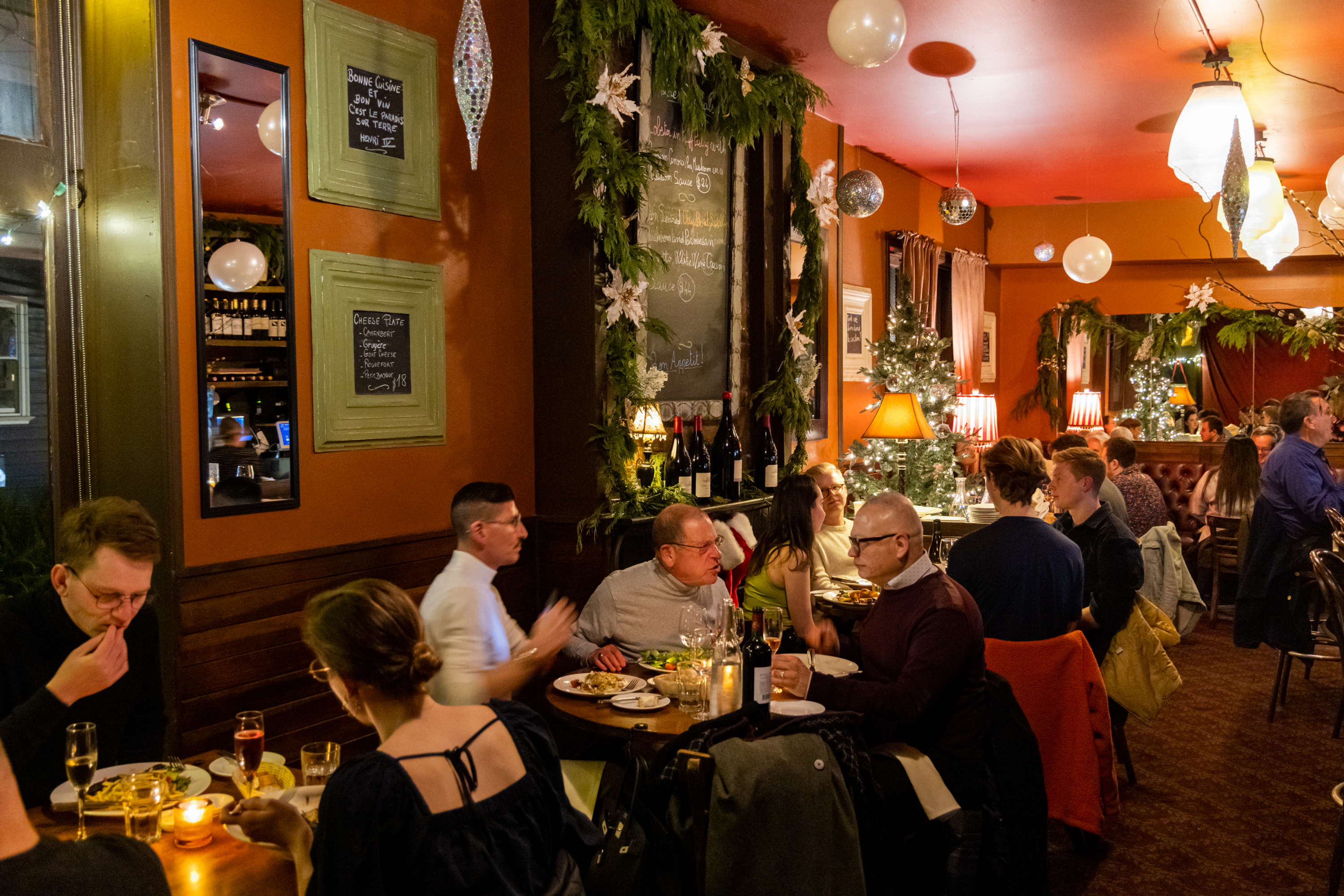 A cozy restaurant filled with diners enjoying meals. The room is warmly lit, decorated with festive greenery, mirrors, and a small Christmas tree.