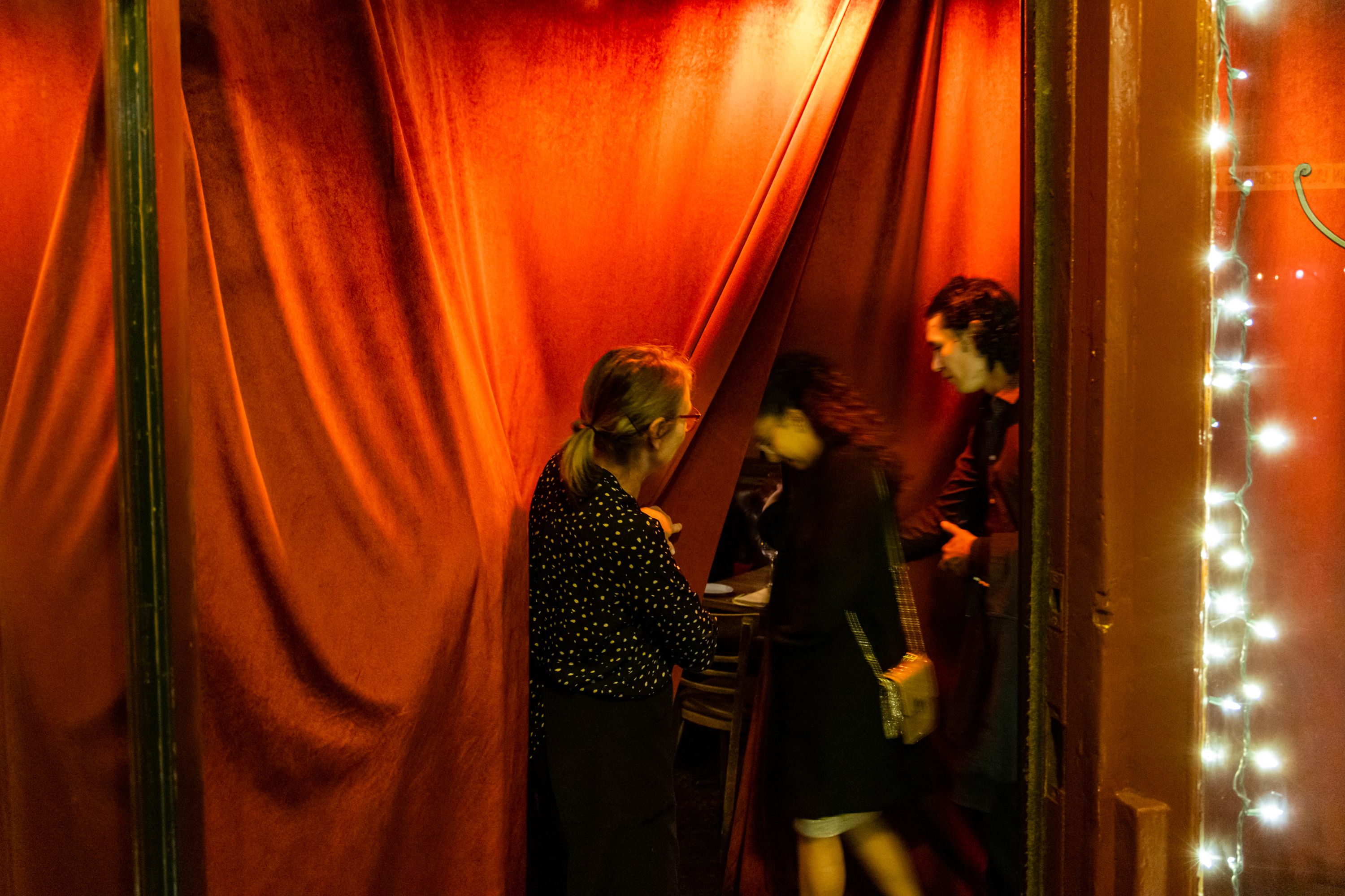 Three people are entering a warmly lit room through red curtains. One person holds the curtain open, while another stands outside. String lights adorn the doorway.