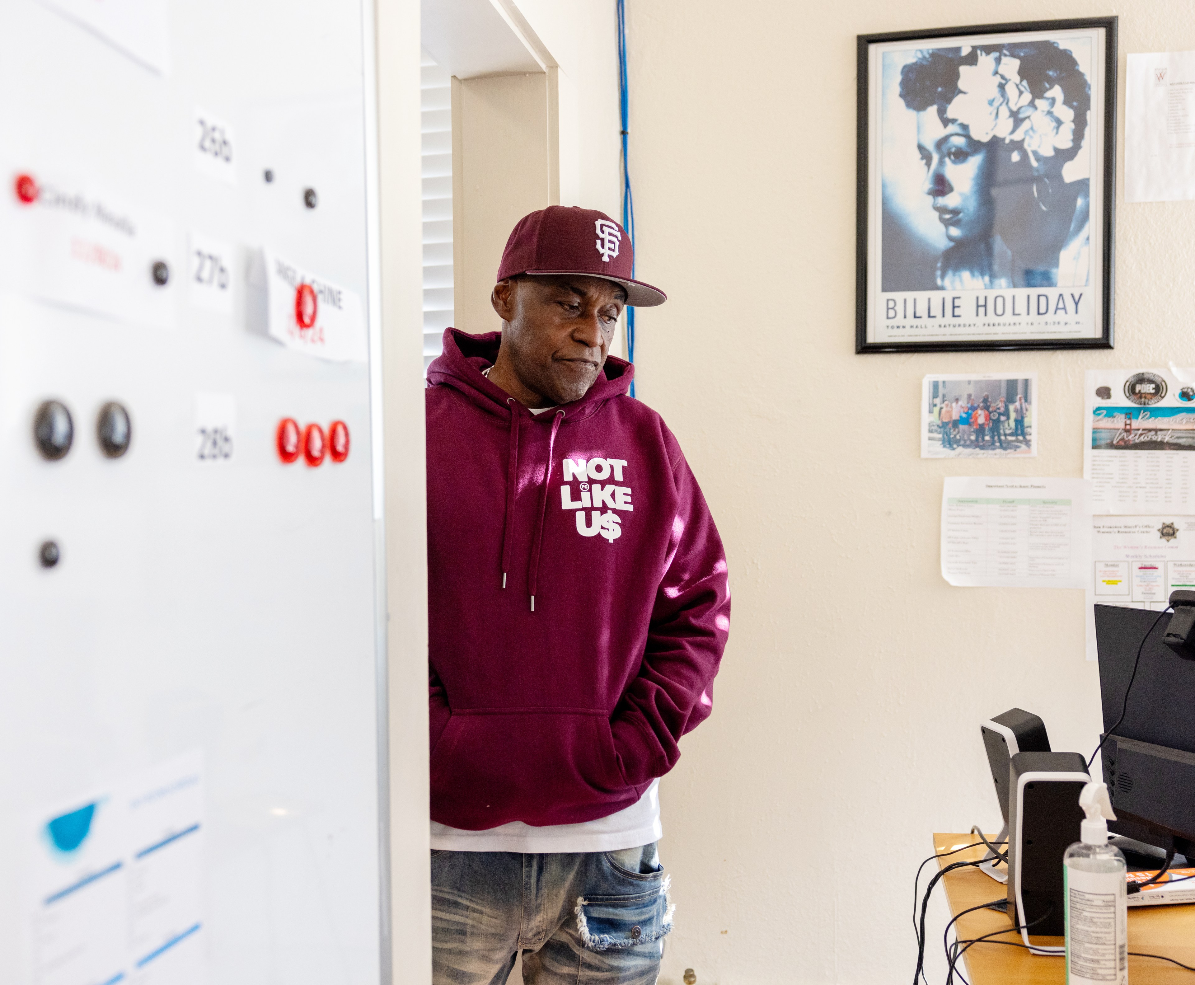 A man in a maroon hoodie and cap stands by a wall with a Billie Holiday poster. A desk with a computer and sanitizer is nearby, along with various notices.