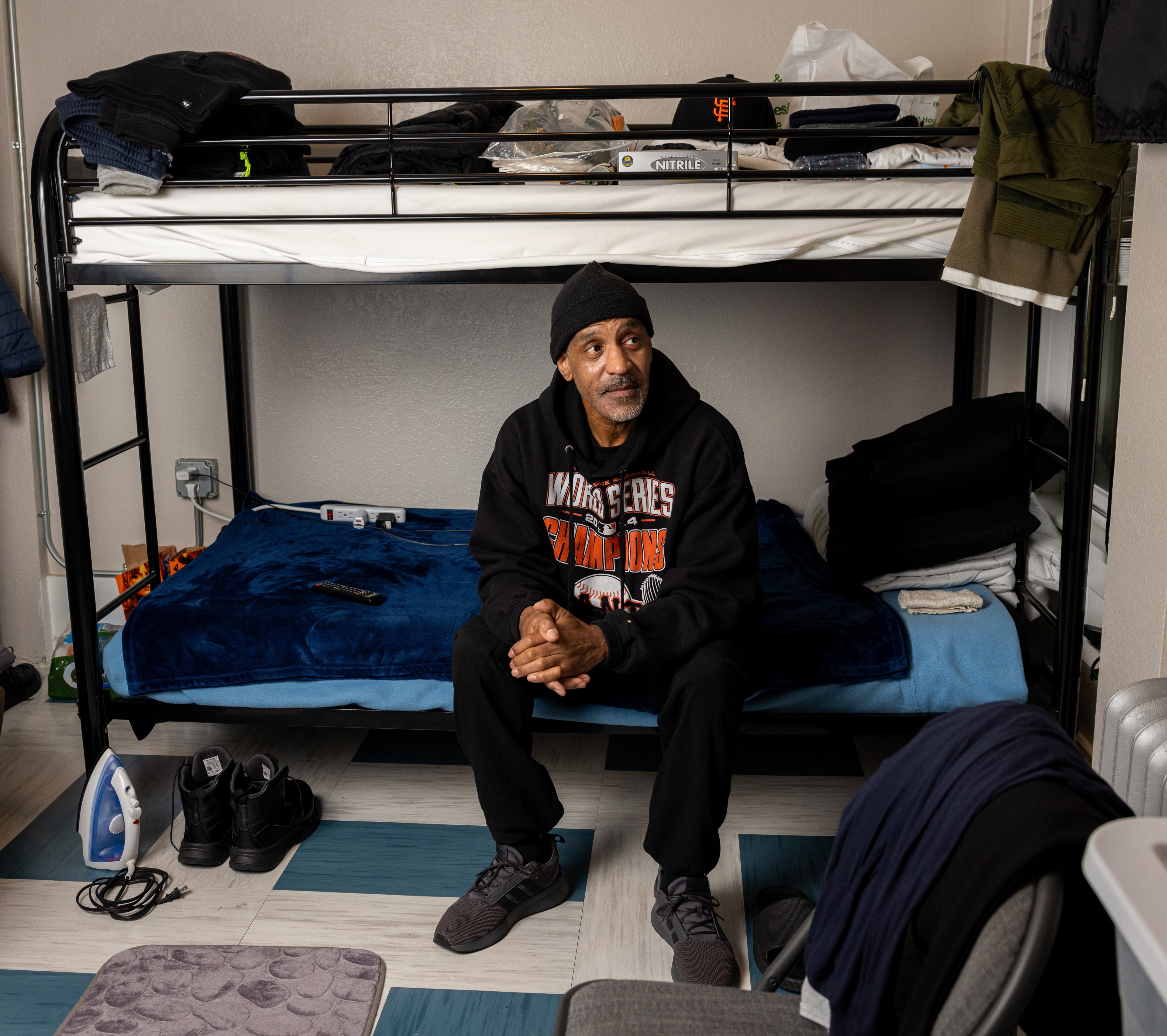 A man sits on the lower bunk of a bed in a room. The bed is covered with a blue blanket, and various items are stored on the top bunk. An iron is on the floor nearby.