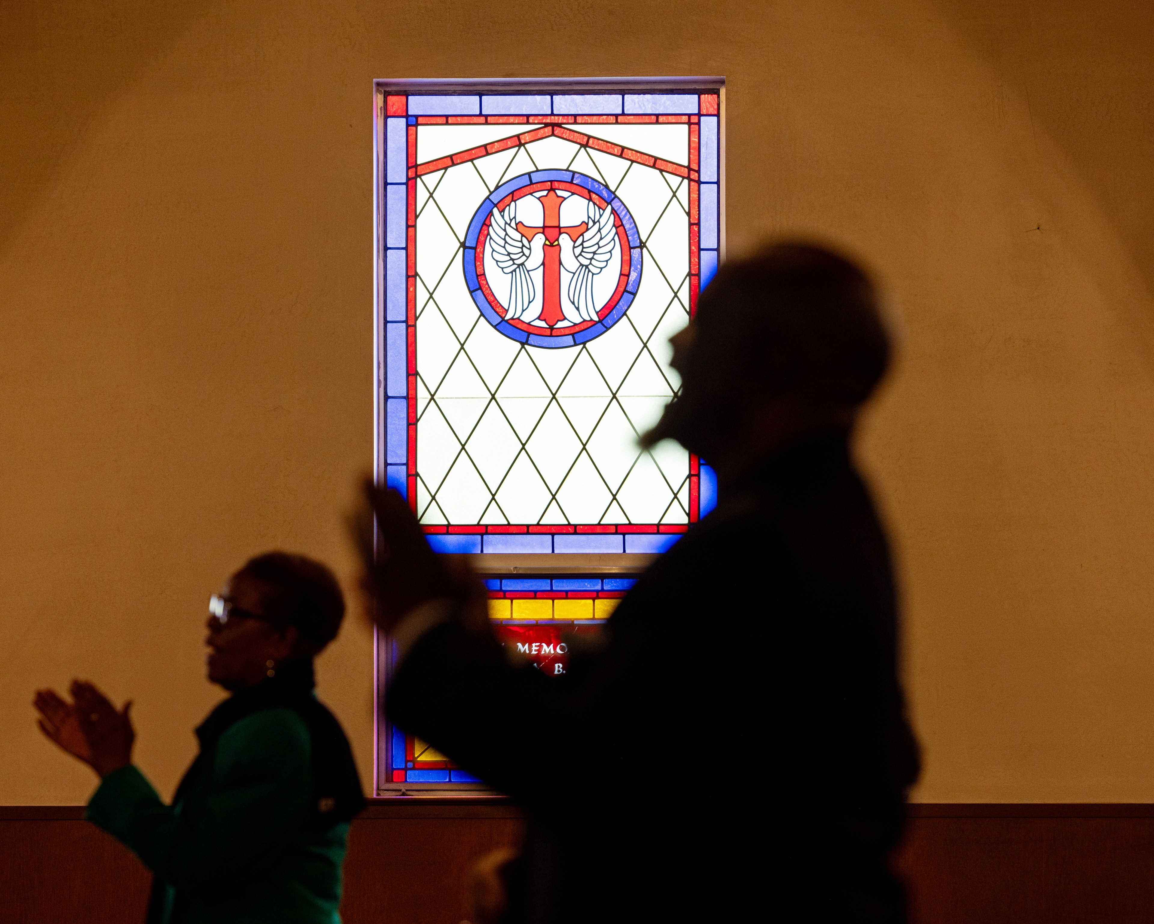 The image shows two silhouetted figures clapping in front of a stained glass window with a red and blue border, featuring a cross with wings.