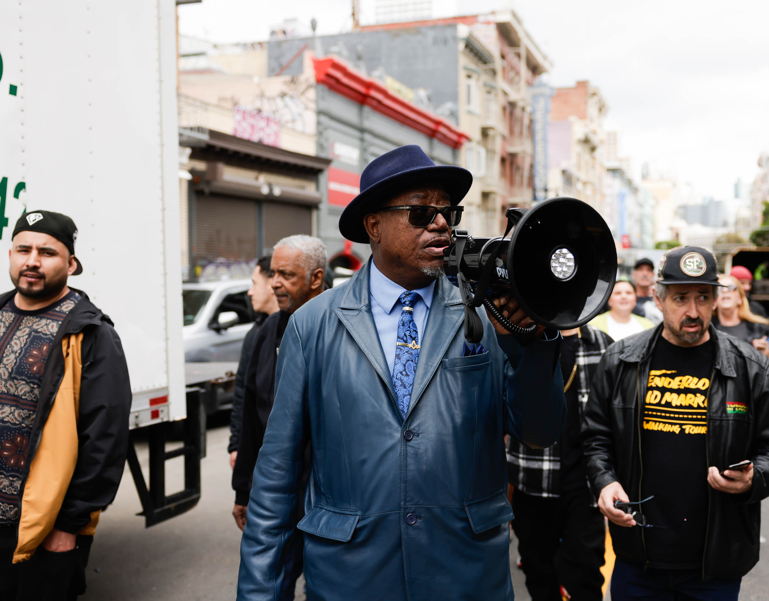 A man in a blue coat and hat holds a megaphone, addressing a group of people on a city street. Other individuals surround him, some looking attentive.