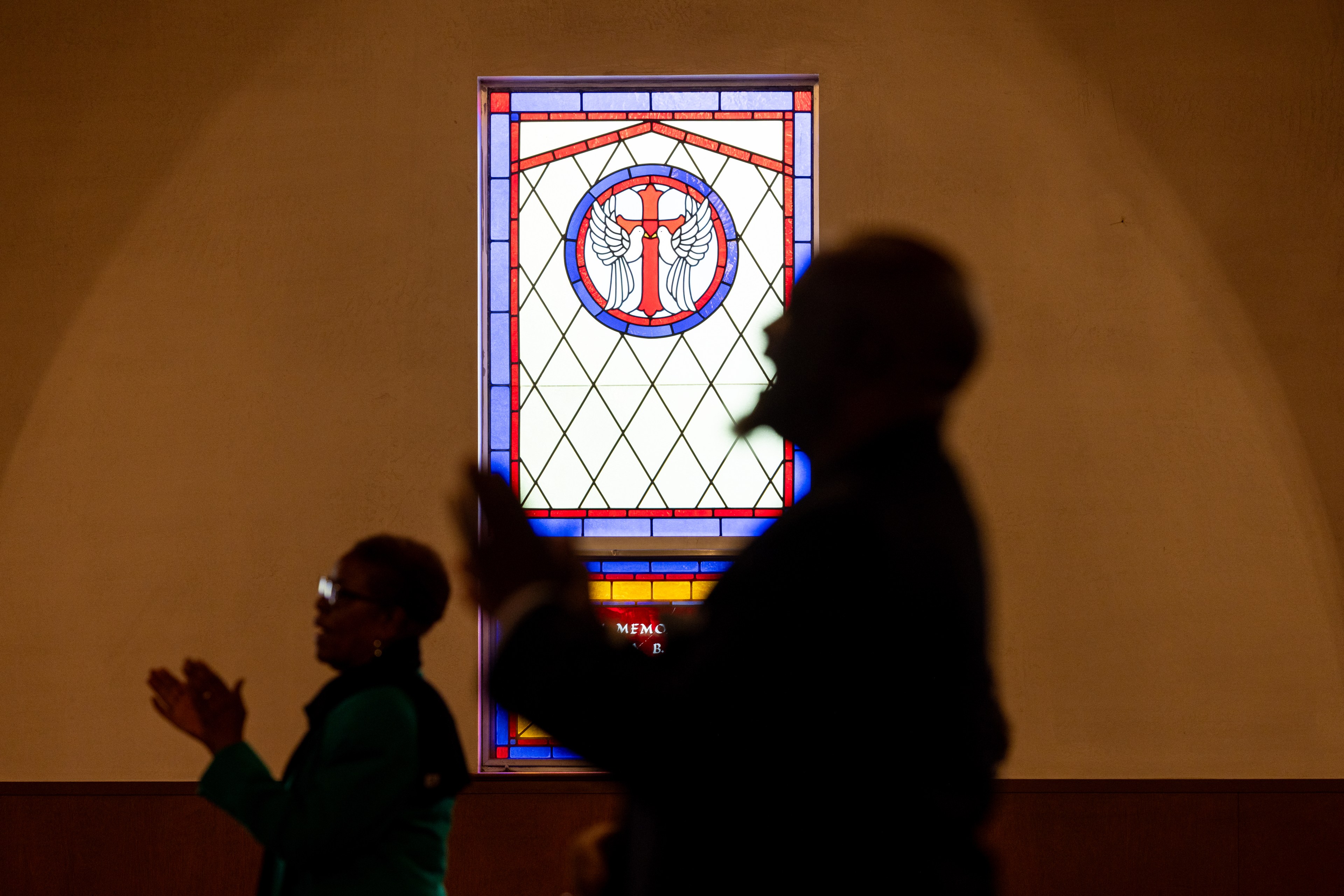 Two silhouetted people are seen clapping near a colorful stained glass window featuring a cross with wings in a church setting.