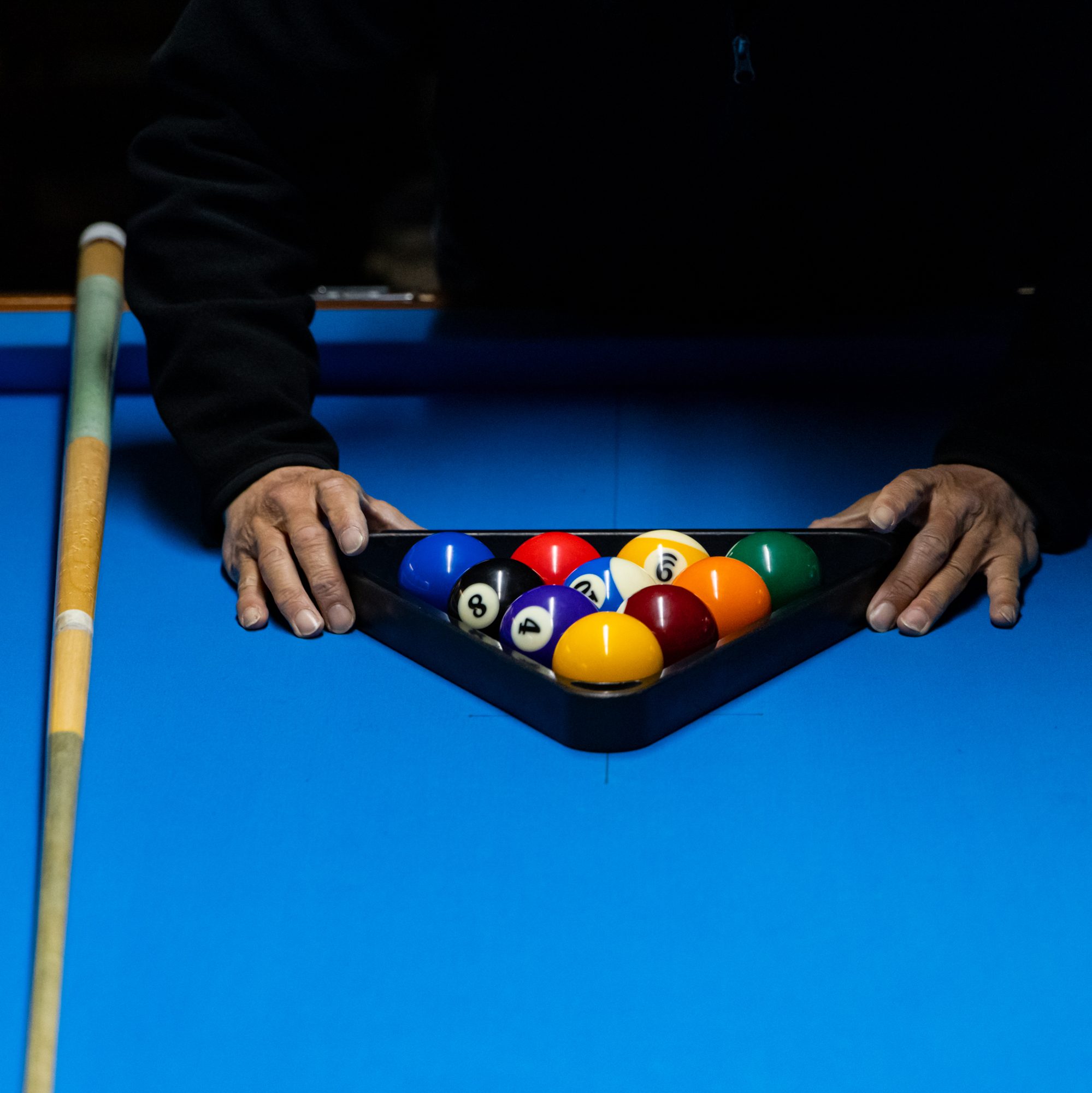 A person sets up colorful billiard balls in a triangular rack on a blue pool table, with a cue stick resting beside them.