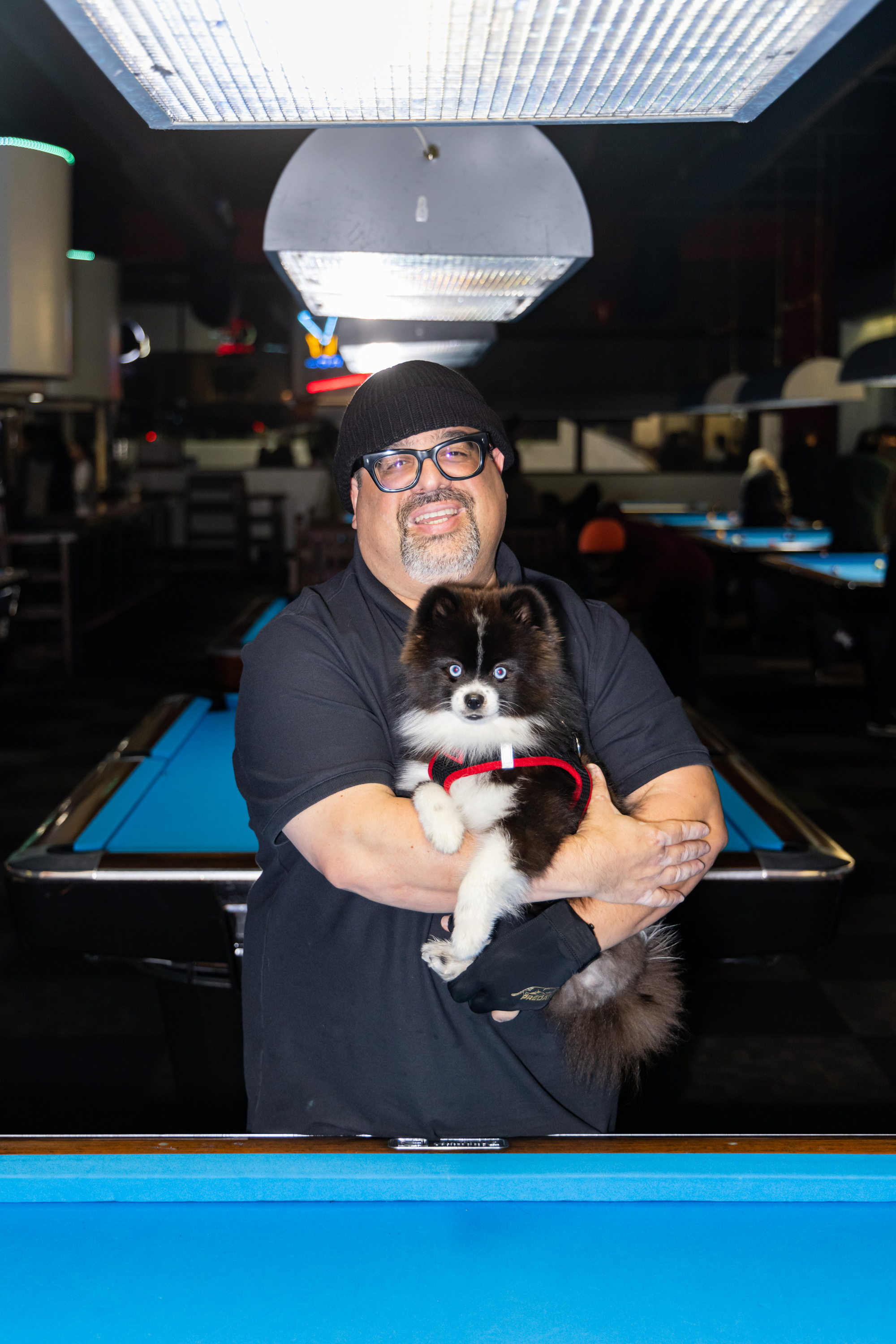 A smiling person with glasses and a beanie holds a small, fluffy black and white dog in their arms. They stand in a pool hall with blue pool tables.