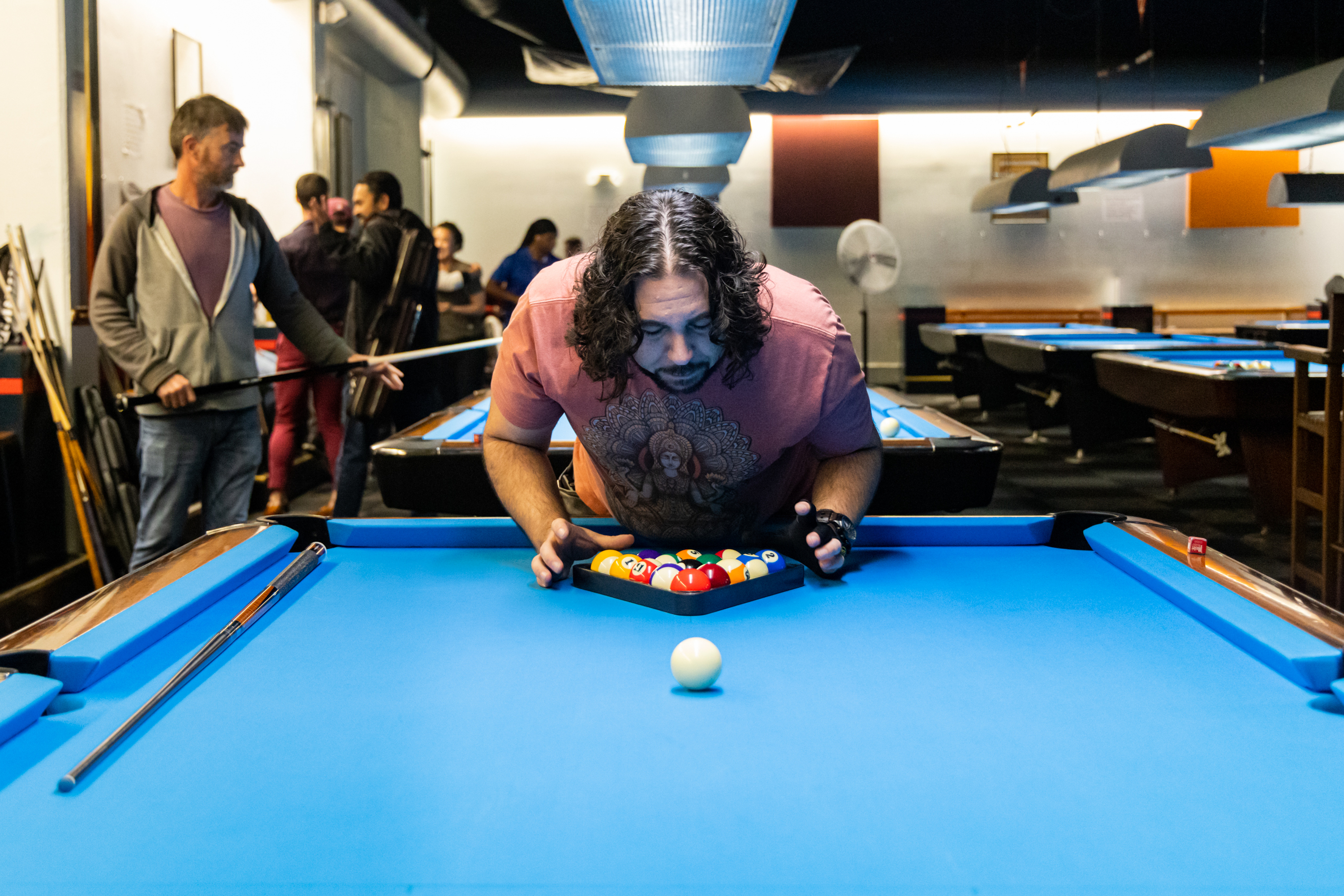 A man with long hair is arranging pool balls on a blue-felted pool table, focusing intently. Several people are in the background, holding pool cues.