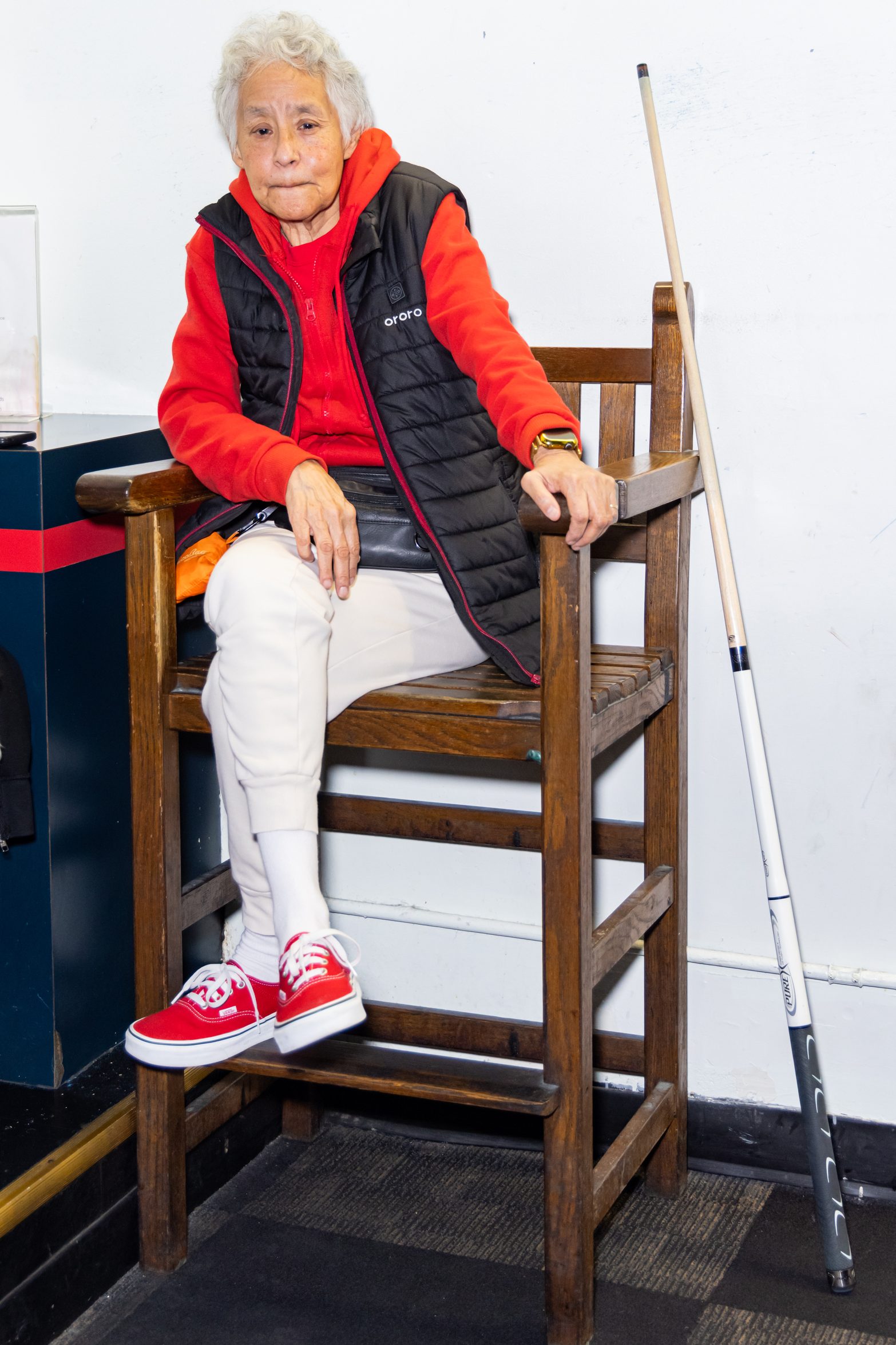 An elderly person in a red hoodie and black vest sits on a wooden chair, with a pool cue beside them. A menu and a coffee cup are nearby.