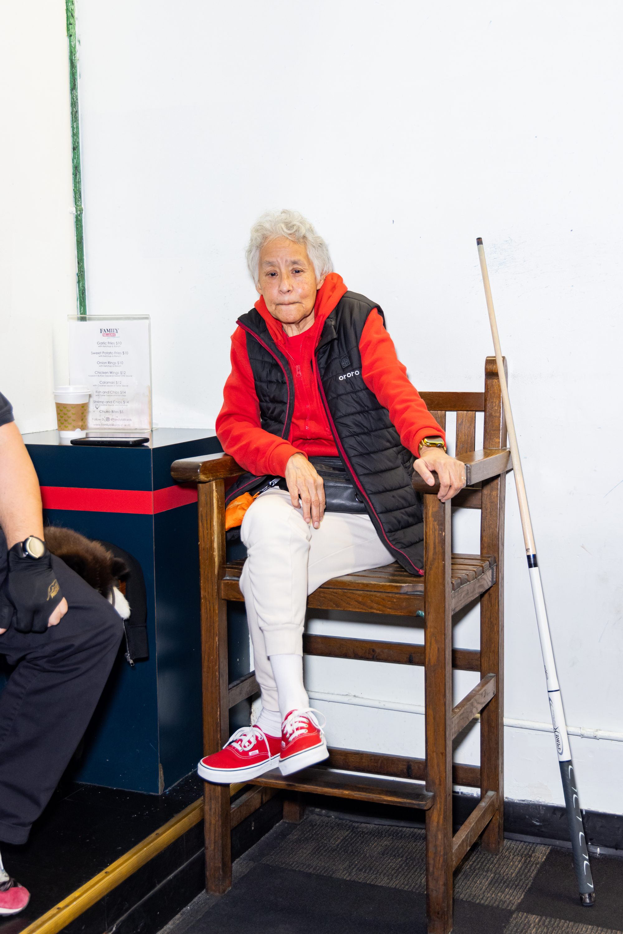 An elderly person in a red hoodie and black vest sits on a wooden chair, with a pool cue beside them. A menu and a coffee cup are nearby.