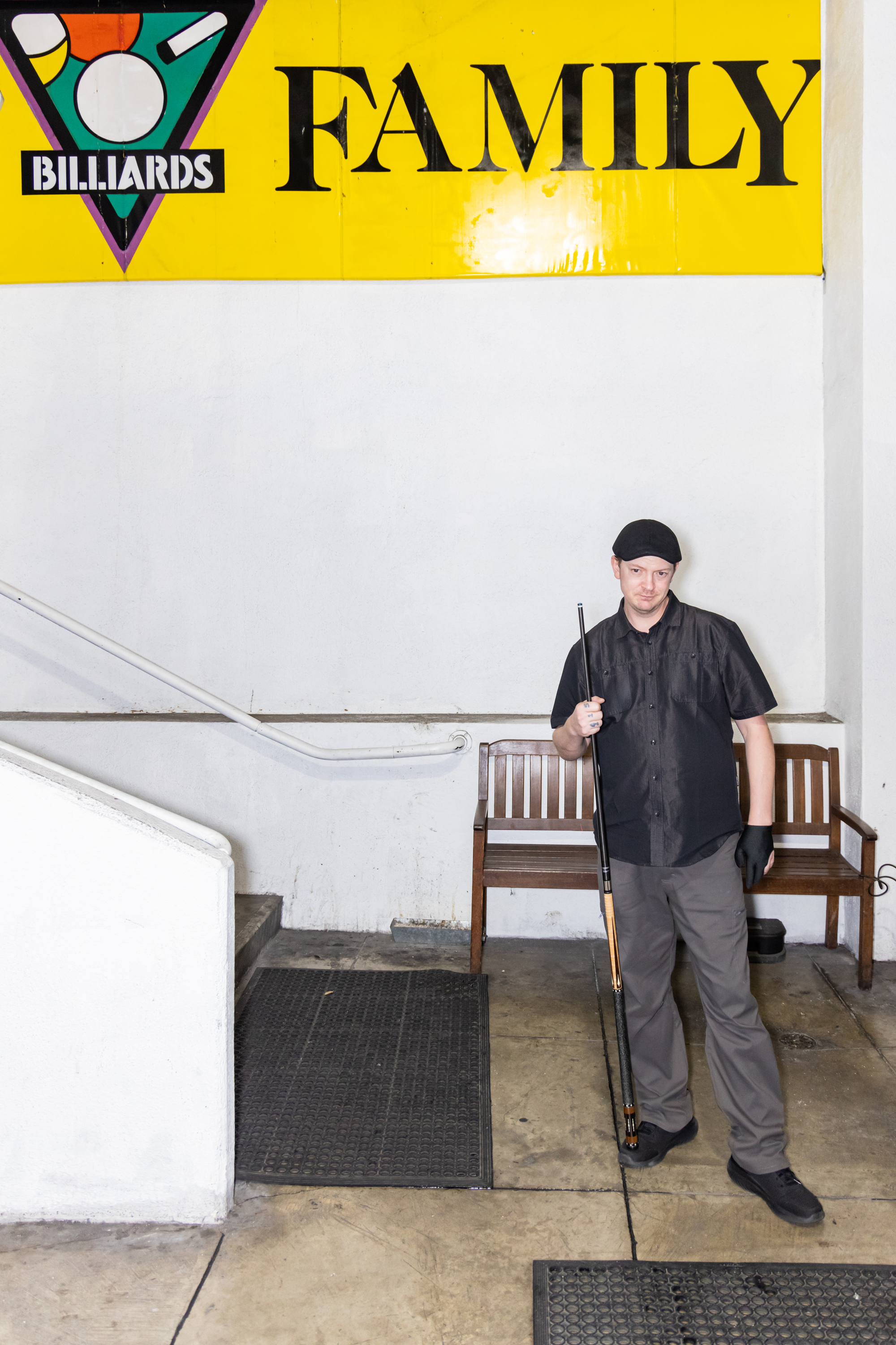 A person stands holding a pool cue, wearing a black shirt and cap, in front of a yellow wall with a &quot;Billiards Family&quot; sign. There's a wooden bench nearby.