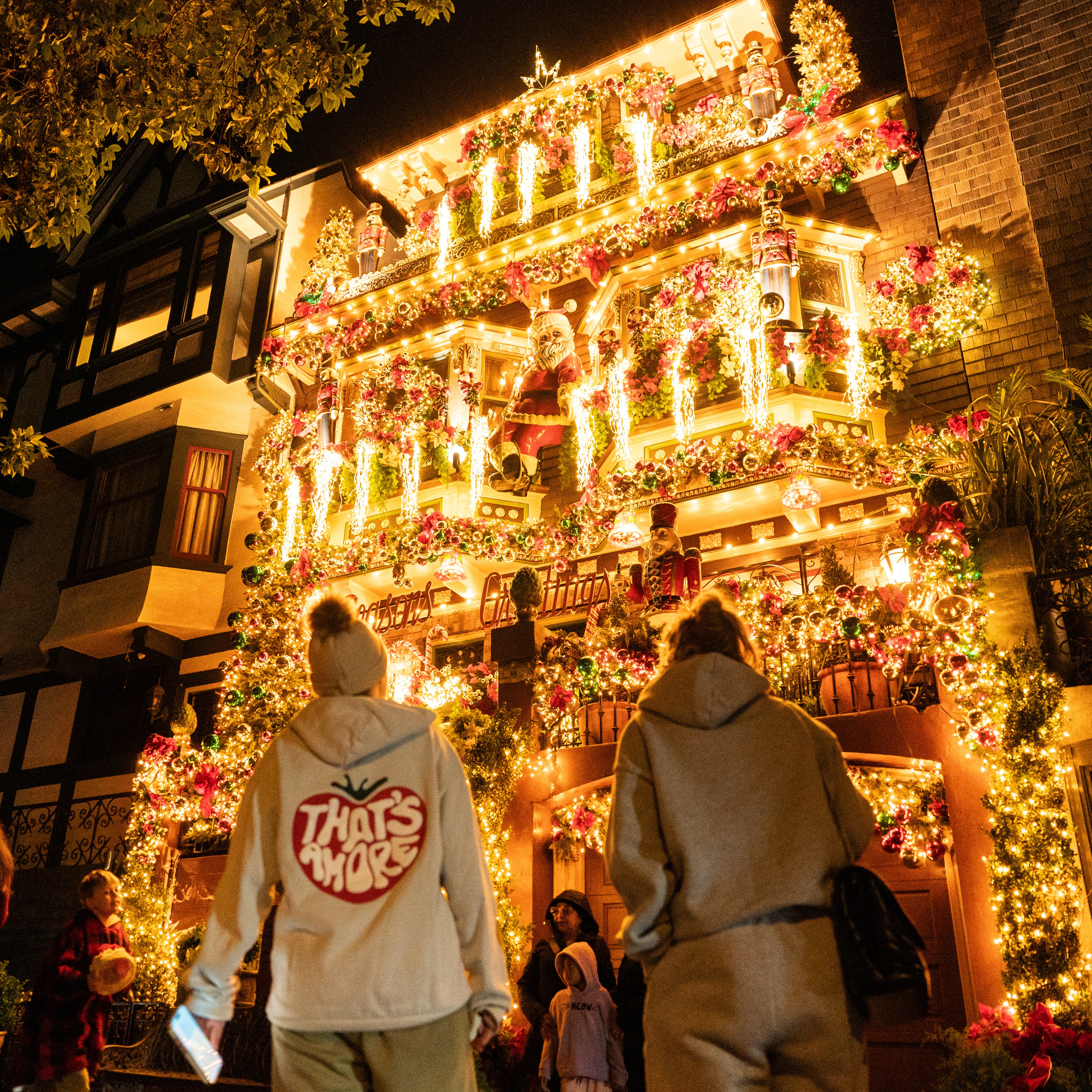 A house is adorned with bright, colorful Christmas lights and decorations. People stand in front, admiring and taking photos as they enjoy the festive display.