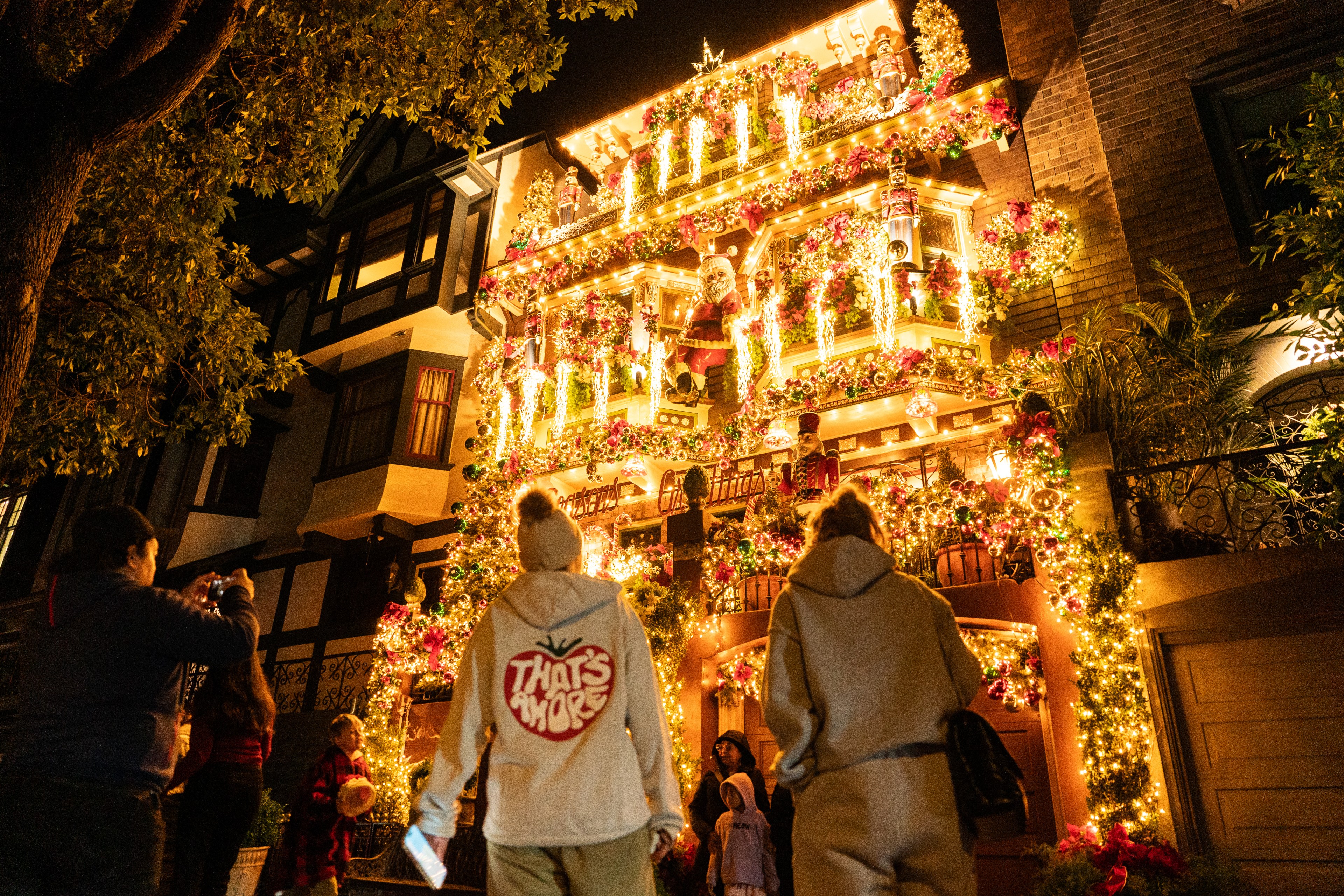 A house is adorned with bright, colorful Christmas lights and decorations. People stand in front, admiring and taking photos as they enjoy the festive display.