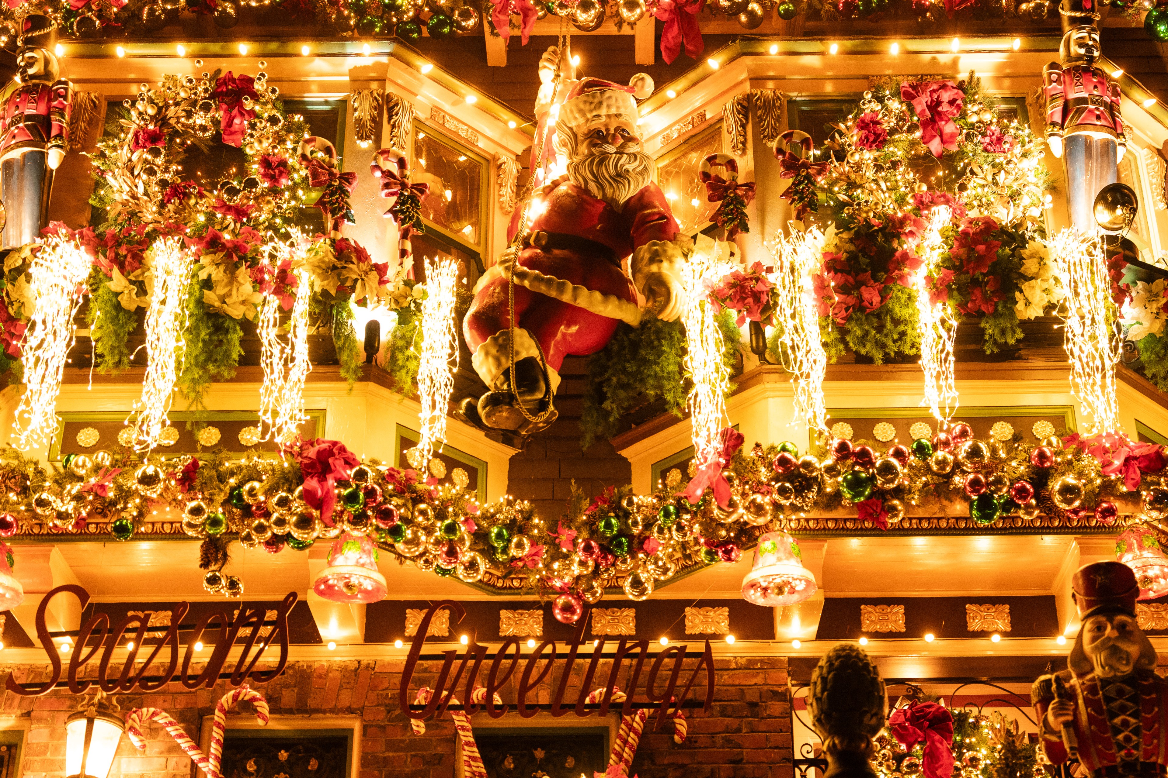 A festive building is adorned with a Santa figure, bright lights, wreaths, garlands, and nutcracker decorations, with &quot;Seasons Greetings&quot; displayed below.