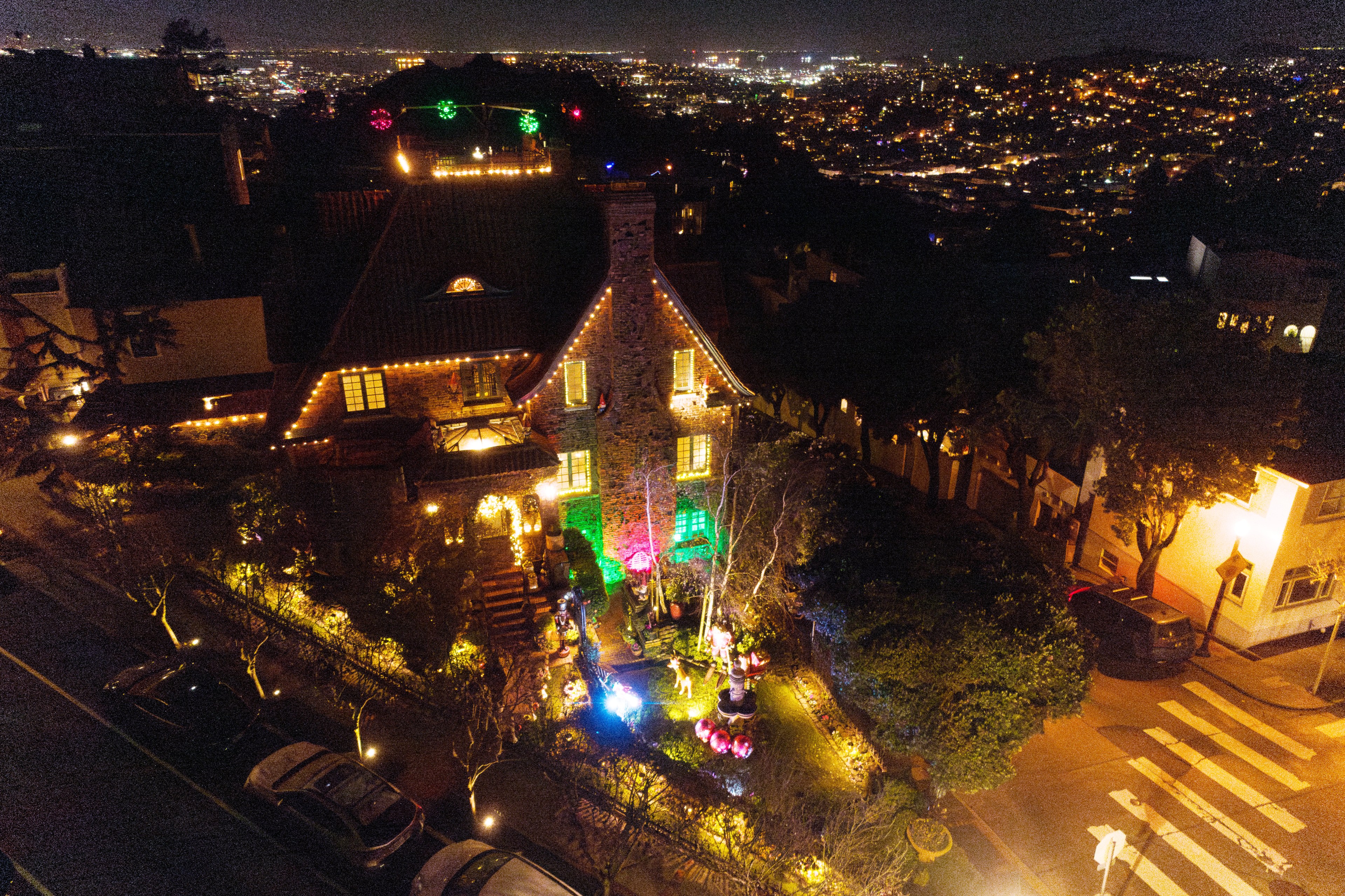 A hillside house adorned with colorful lights stands among trees at night, overlooking a brightly lit cityscape. The garden glows with festive decorations.