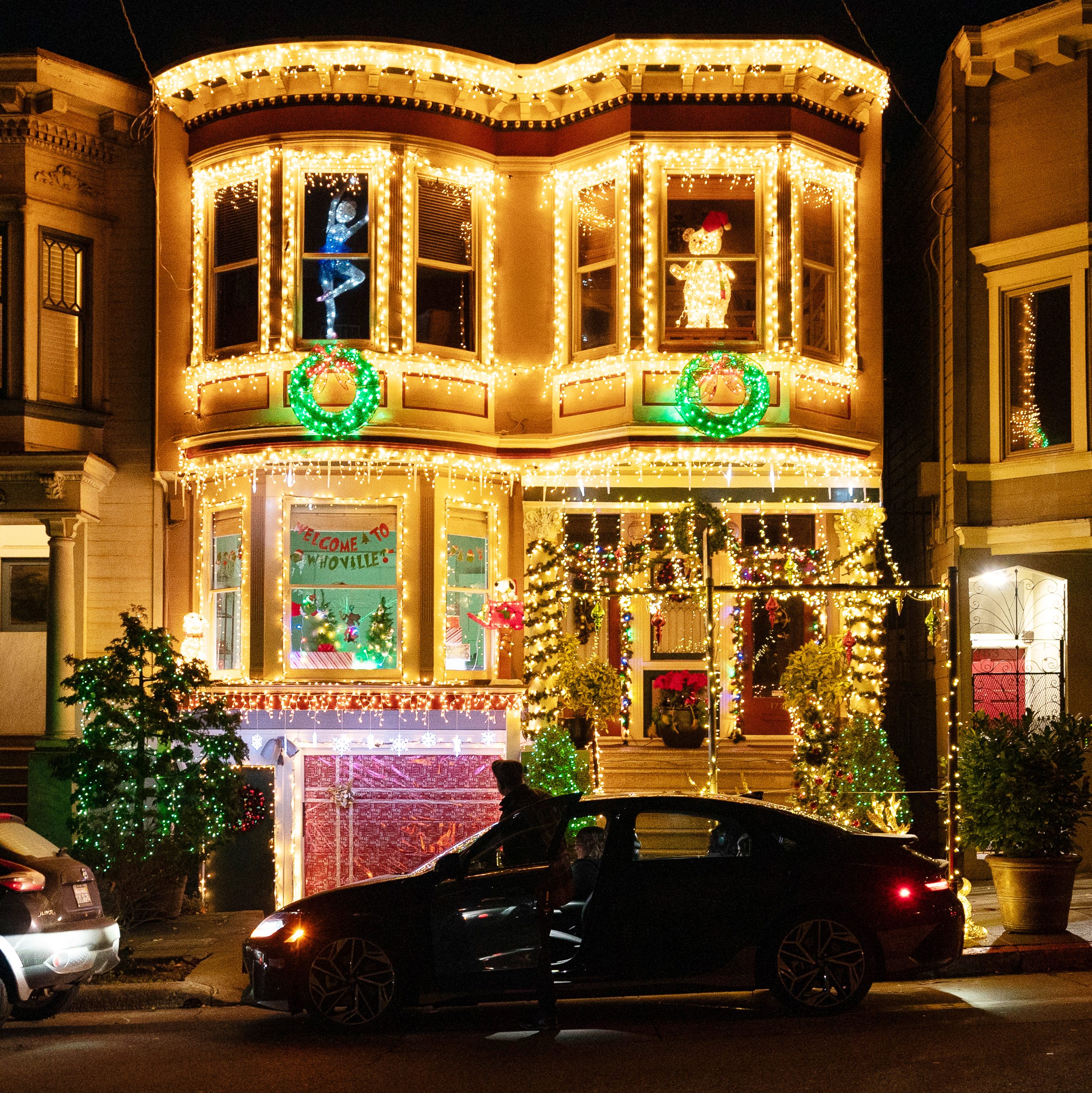 A brightly lit house is decorated with festive lights and wreaths, featuring a &quot;Welcome to Whoville&quot; sign. A person stands beside an open car door in front.