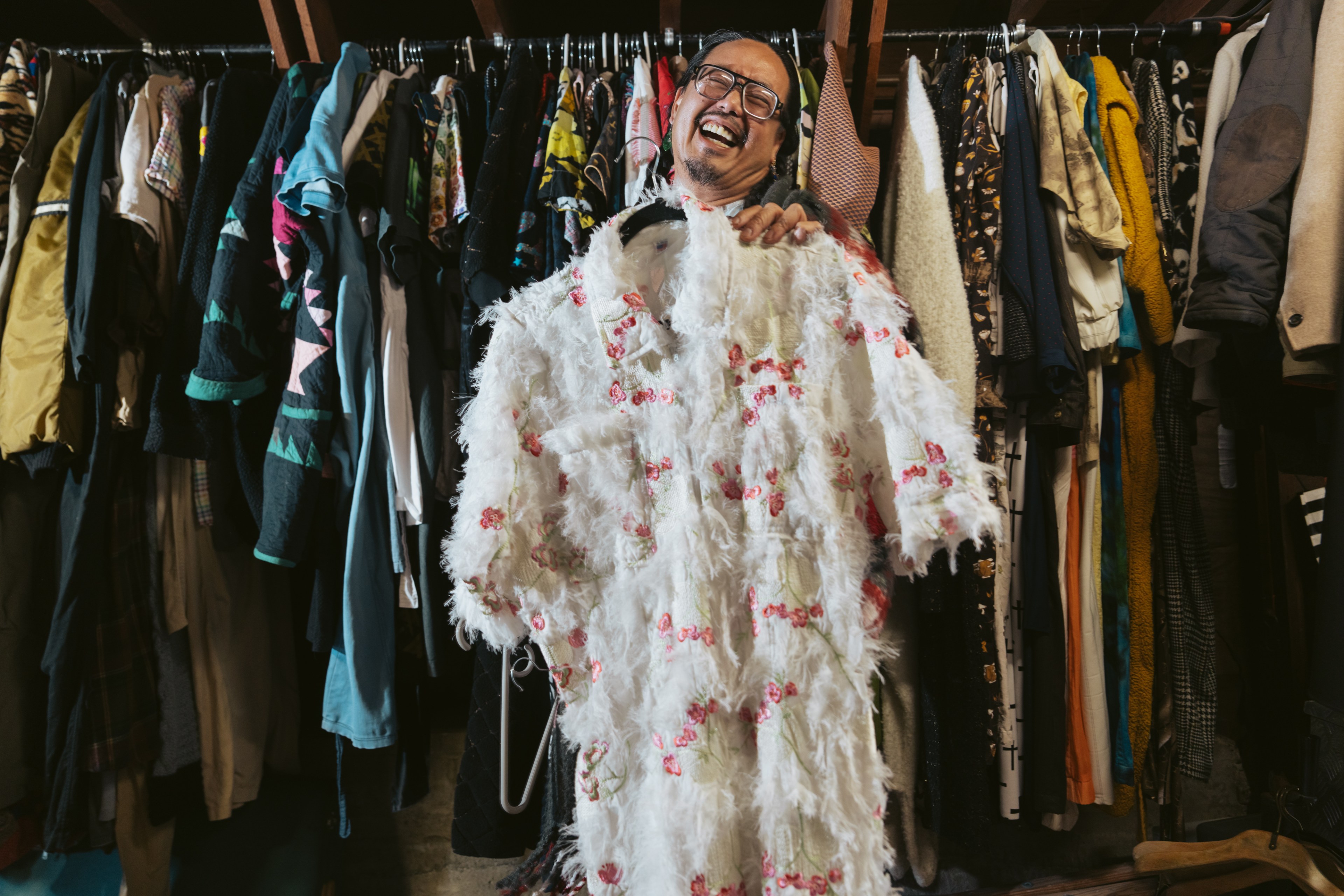 A person smiling broadly holds a fluffy white coat with pink flowers, standing in front of a colorful and diverse clothing rack.