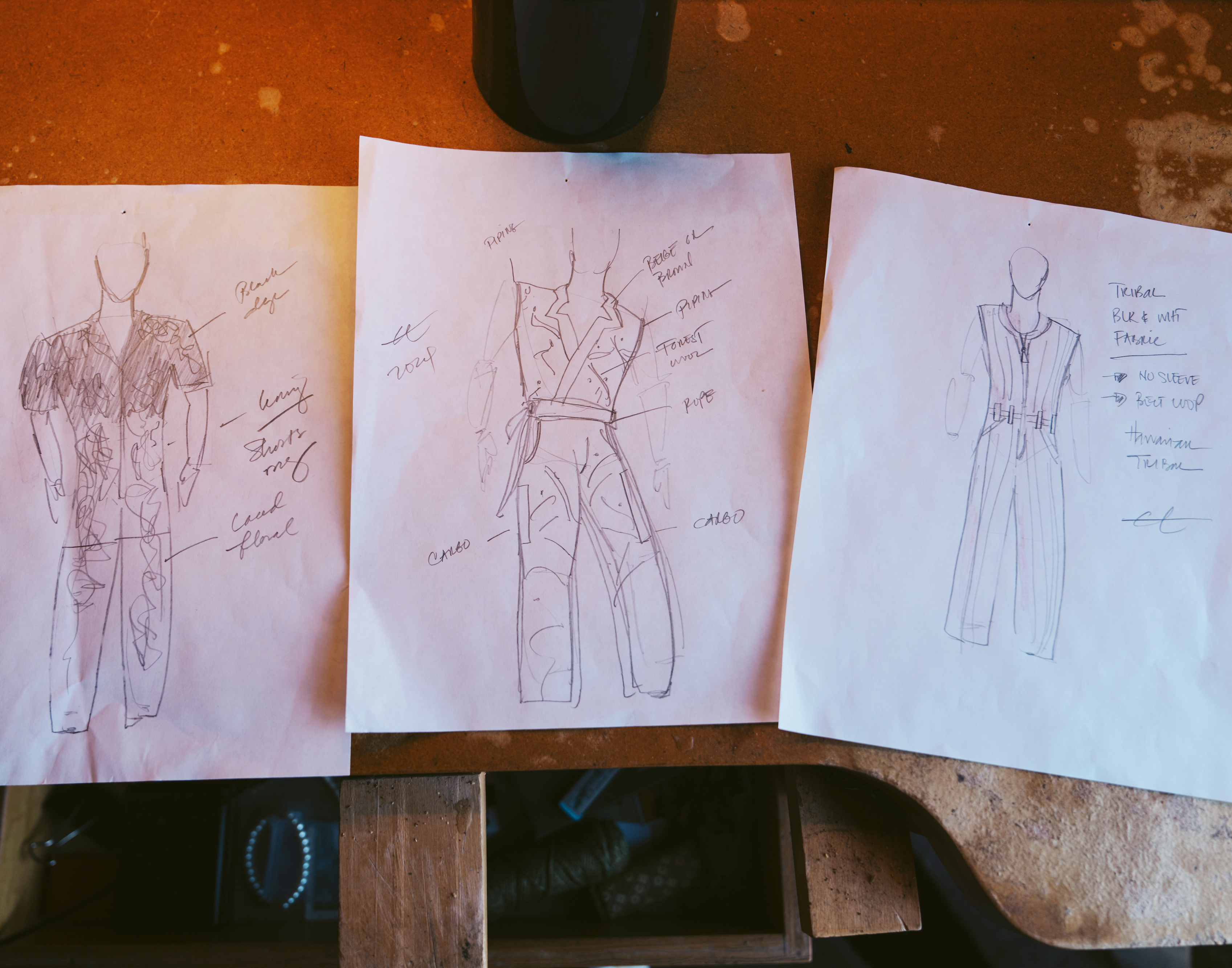 The image shows three hand-drawn fashion sketches on paper pinned to a surface, each depicting a different outfit with notes describing design details.