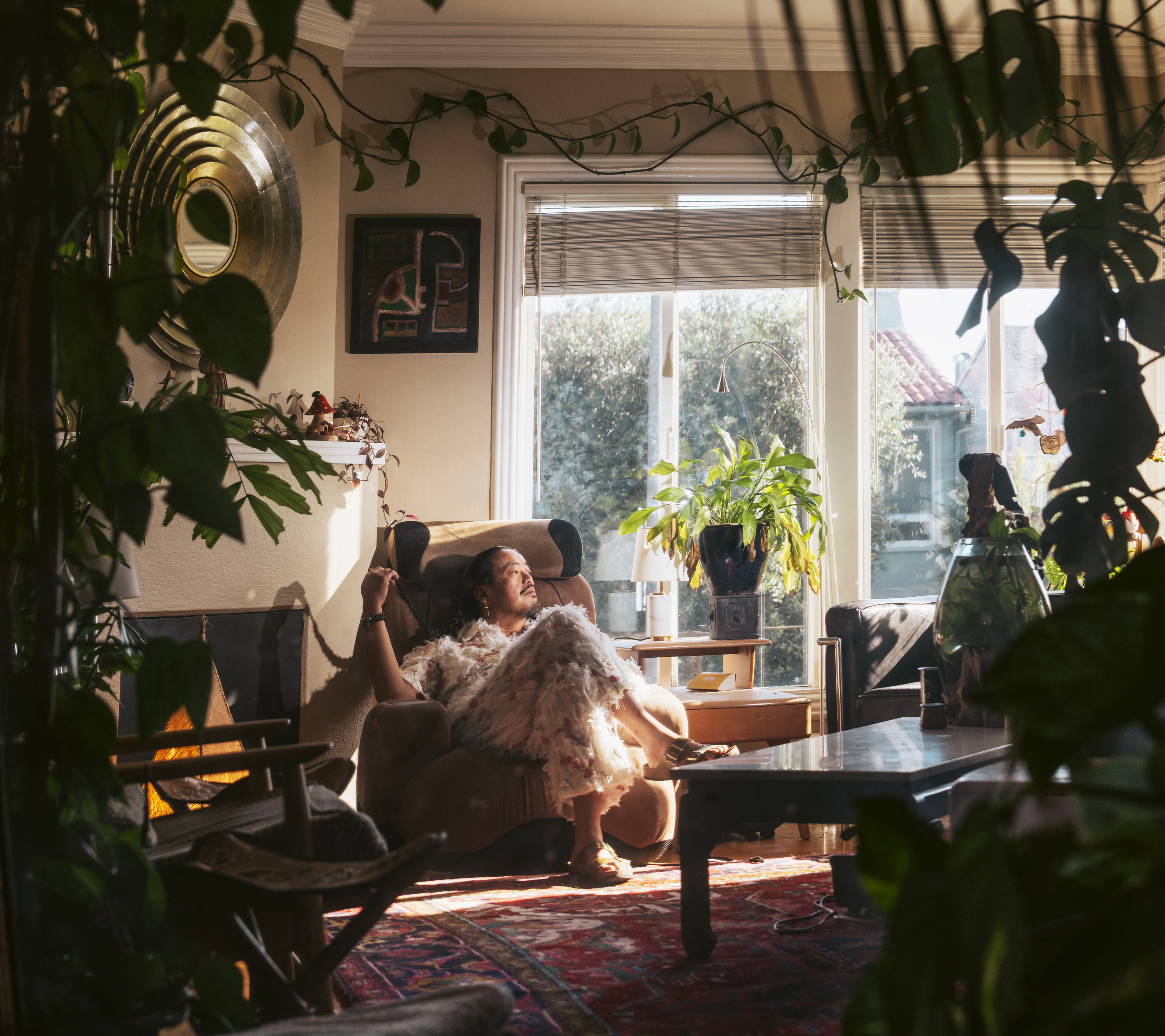 A person relaxes in a cozy, plant-filled room, sitting in an armchair. Soft sunlight streams through large windows, casting gentle shadows on the walls.