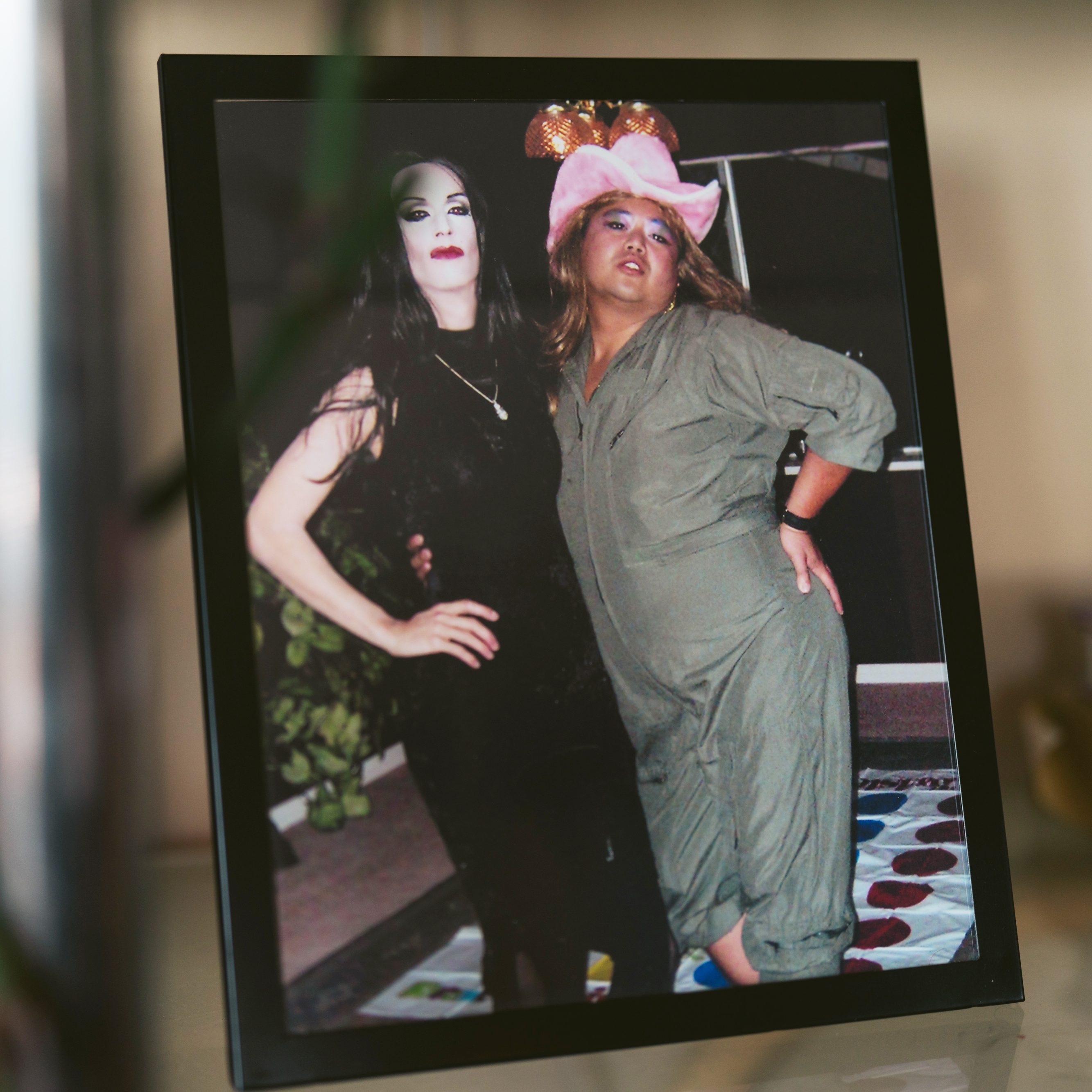 A framed photo shows two people posing playfully. One wears a black outfit with dramatic makeup, and the other sports a pink cowboy hat and a casual jumpsuit.