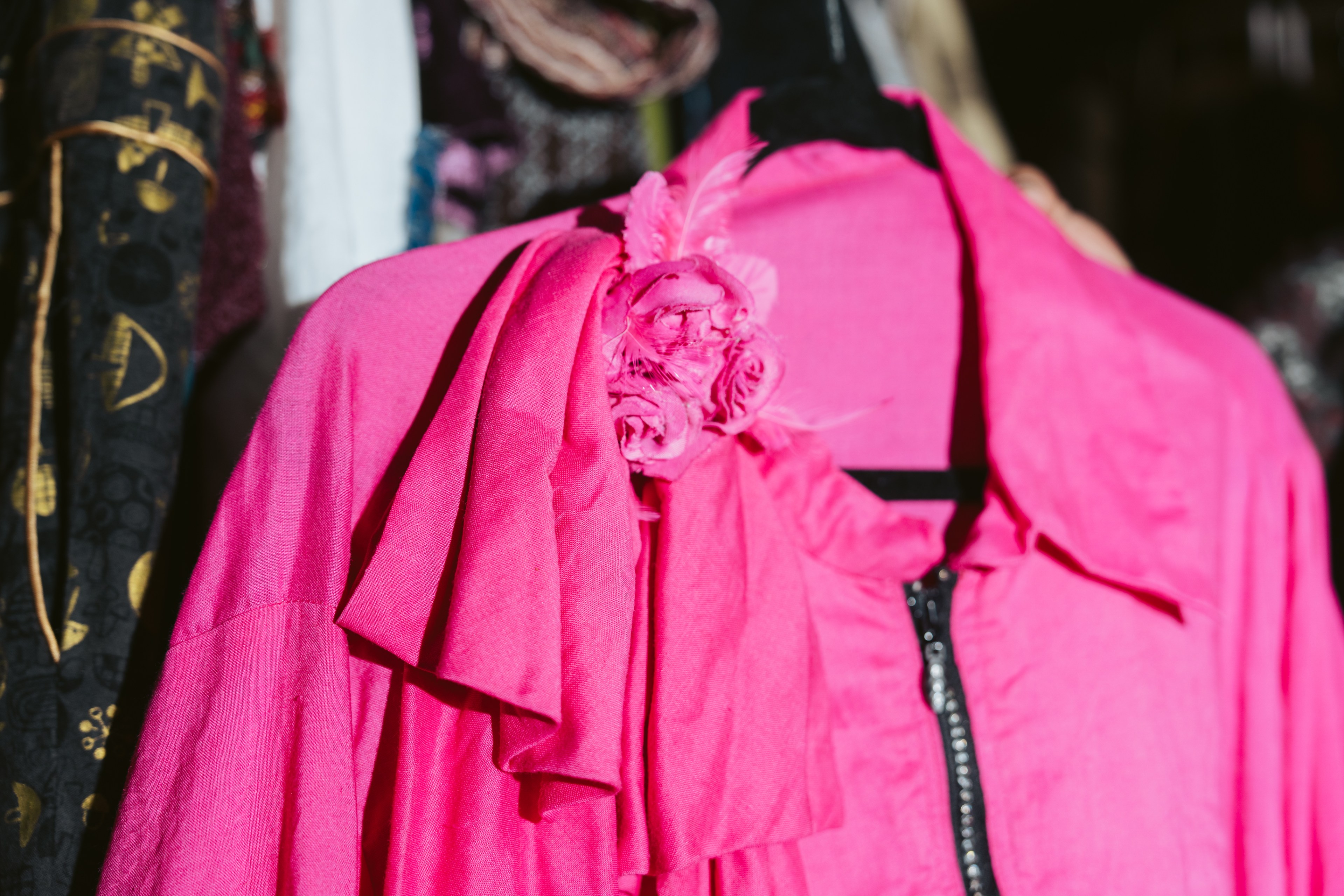 A bright pink blazer is adorned with a matching fabric flower on the shoulder, hung on a black hanger. It contrasts with darker patterned clothes nearby.