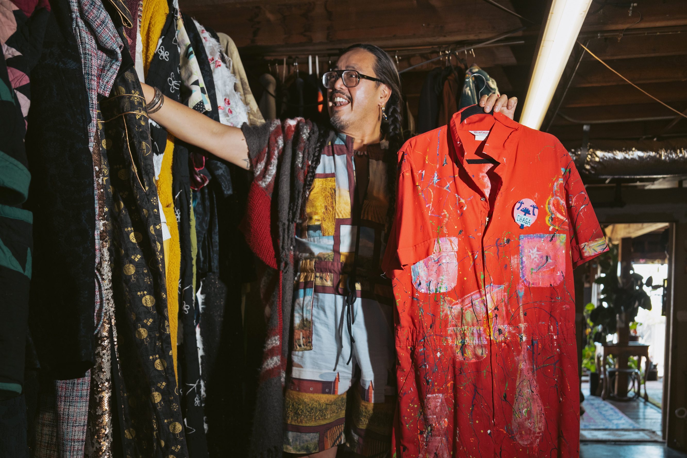 A person stands in a room filled with colorful clothing on racks. They are holding a vivid red jumpsuit splattered with paint. The space is warmly lit and eclectic.