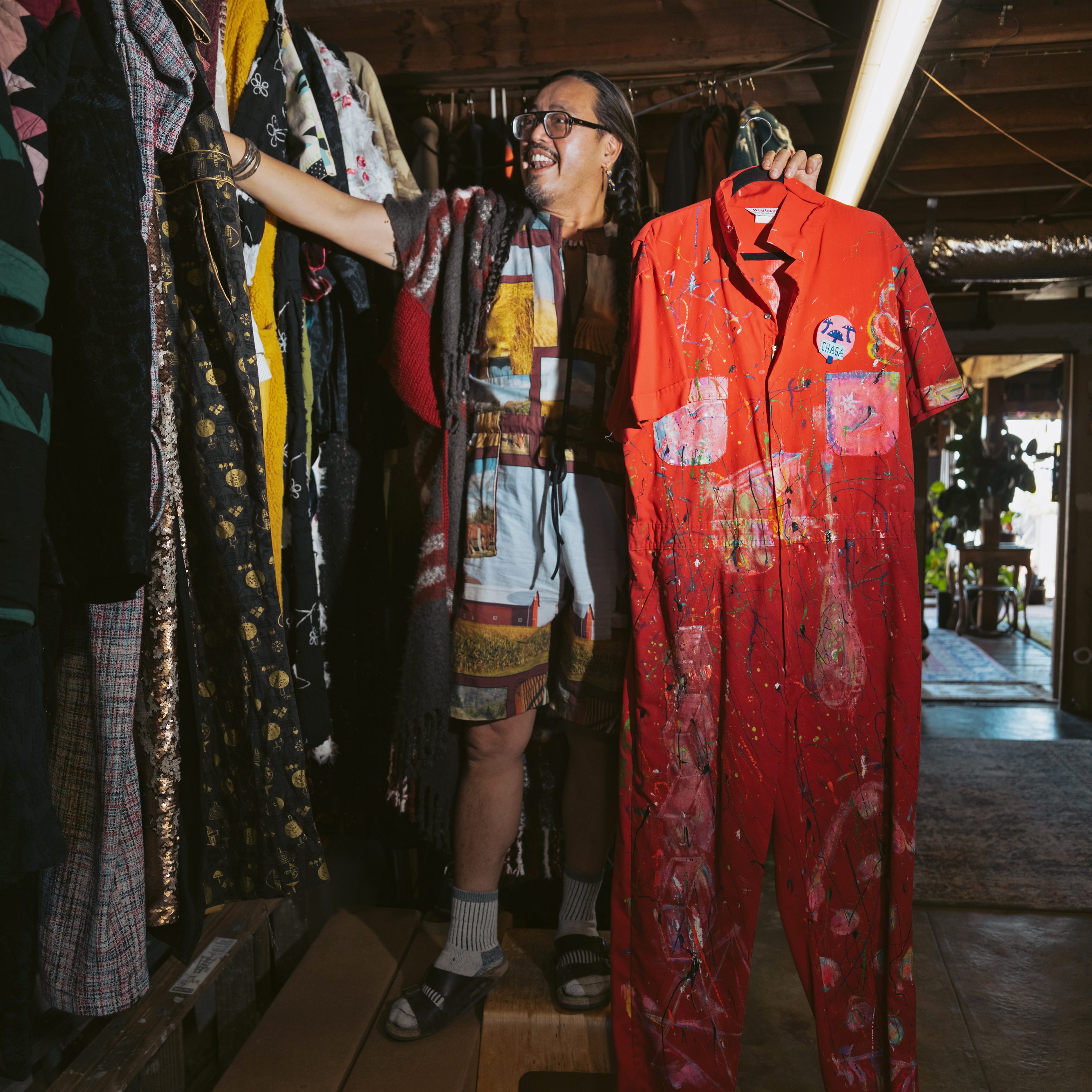 A person stands in a room filled with colorful clothing on racks. They are holding a vivid red jumpsuit splattered with paint. The space is warmly lit and eclectic.