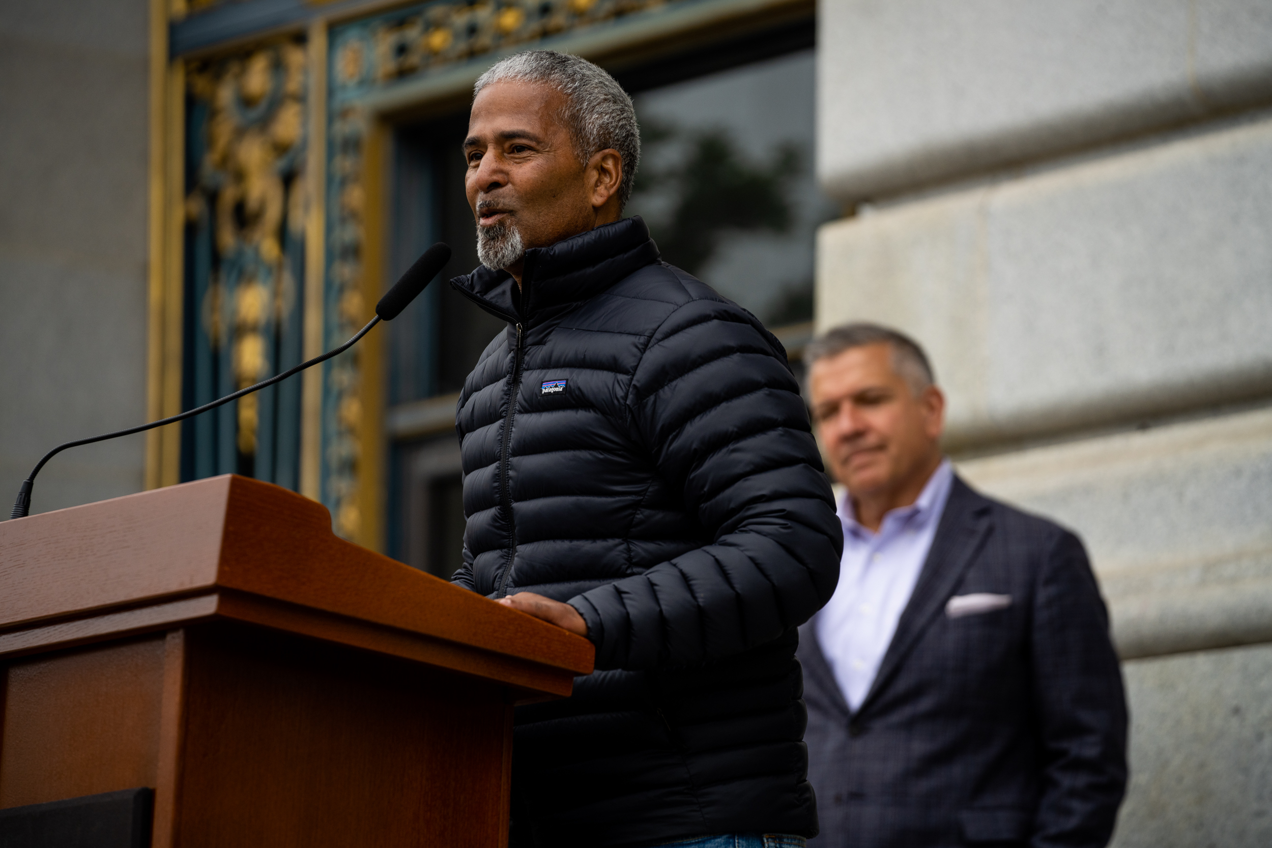 A man in a black puffer jacket speaks at a podium with a microphone. Another man stands in the background, wearing a blazer. The setting appears formal.