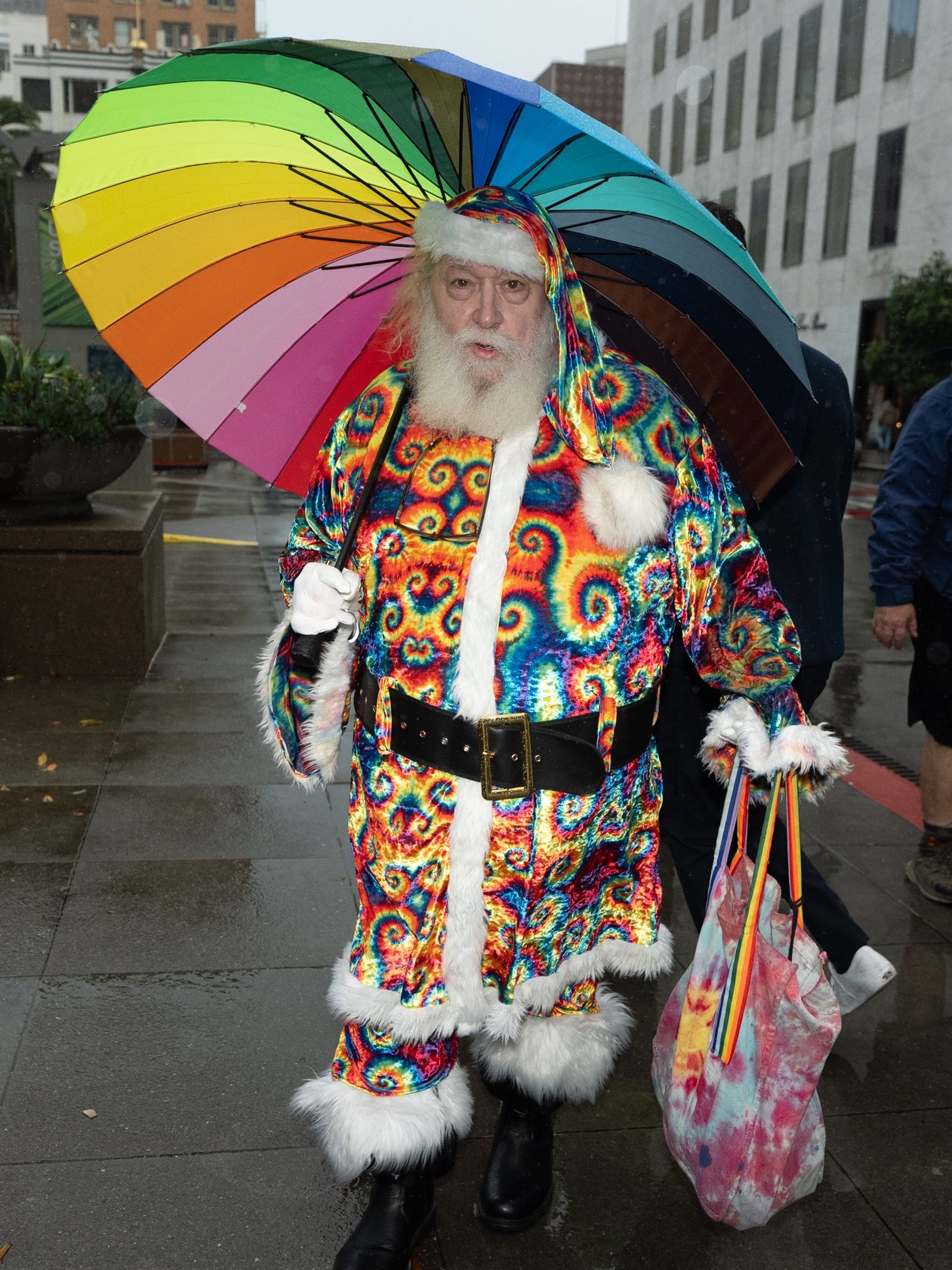 A person in a colorful, patterned Santa suit holds a vibrant rainbow umbrella and carries a tie-dye bag, walking on a wet, downtown city sidewalk.