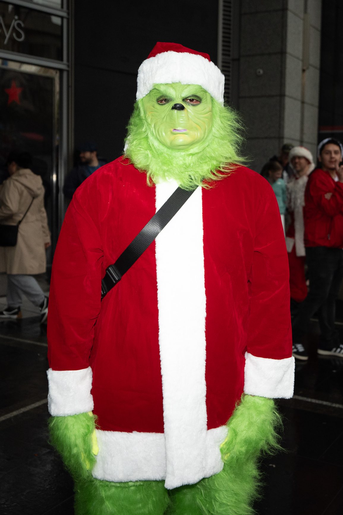 A person is dressed as the Grinch, wearing a green mask and a red Santa suit with white trim, standing on a city street.