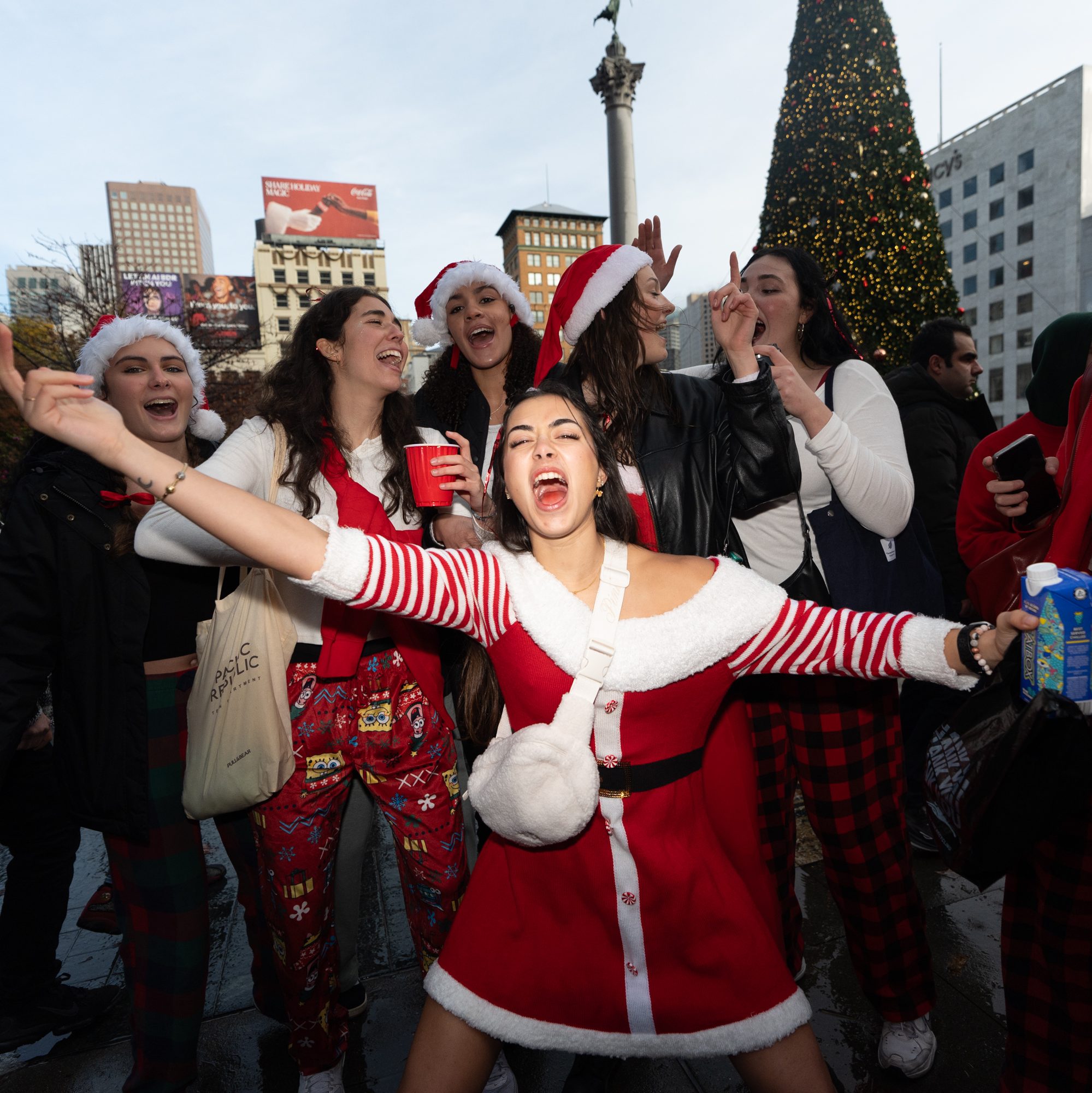 A joyful group of people, mostly women wearing festive Santa hats and red outfits, are celebrating outdoors near a decorated Christmas tree.