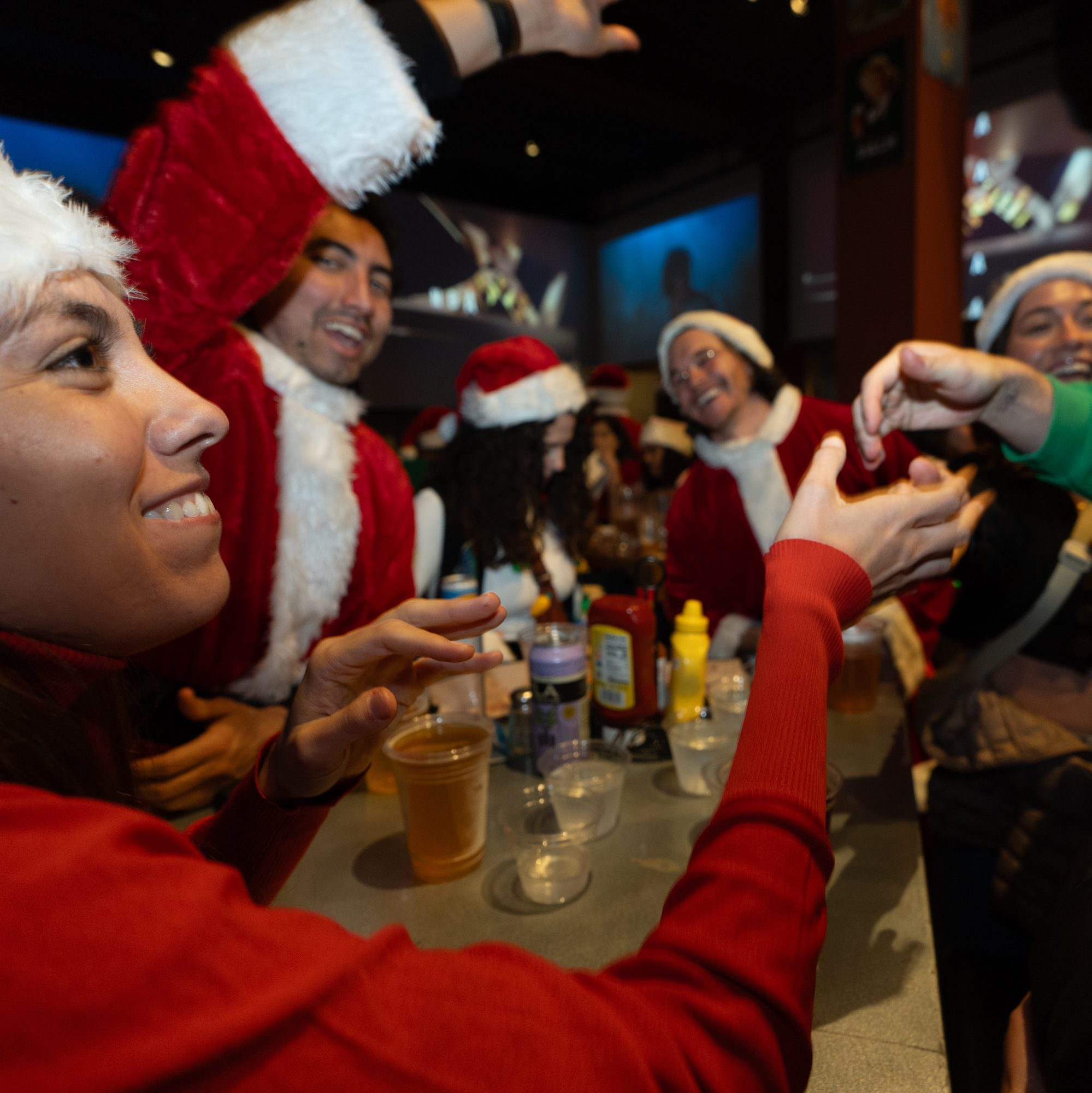 People in Santa hats and festive attire are joyfully gathered around a table with drinks and condiments, smiling and interacting in a lively atmosphere.