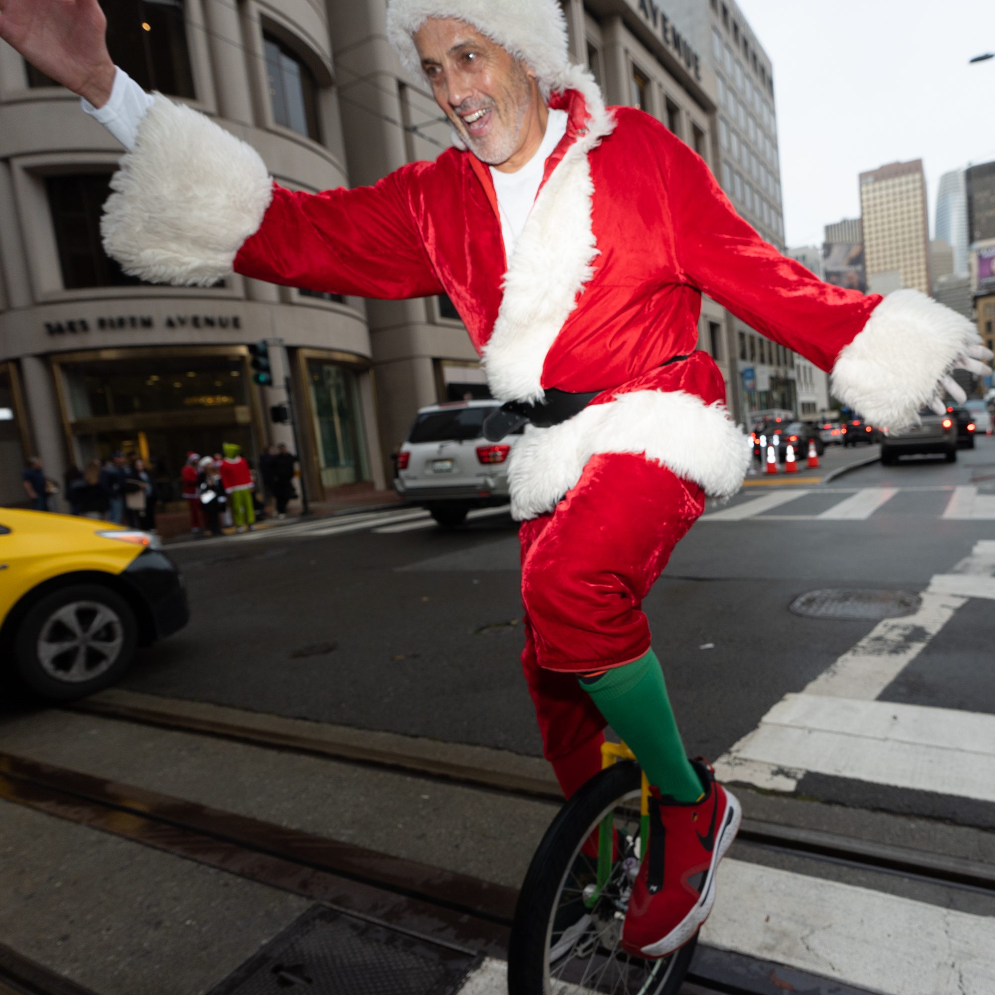 A person dressed as Santa rides a unicycle on a city street, smiling and waving. There are cars, pedestrians, and tall buildings in the background.