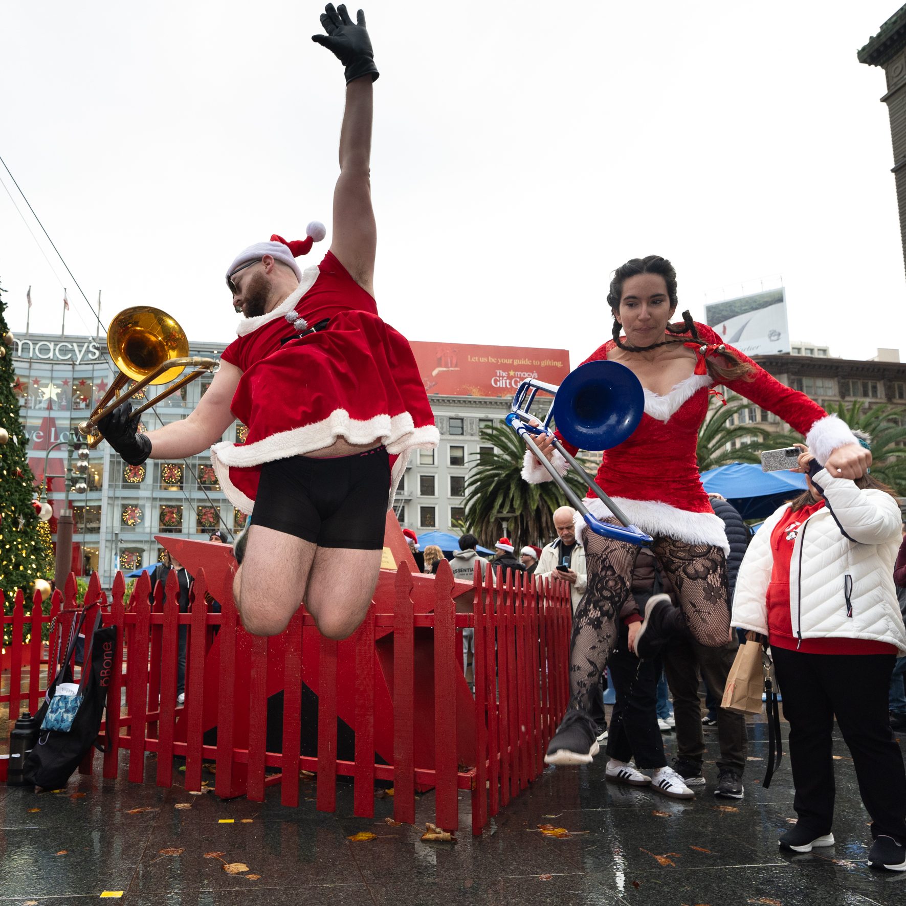 People in festive outfits, including Santa hats, play instruments near a large Christmas tree. One person jumps energetically, while others watch and smile.