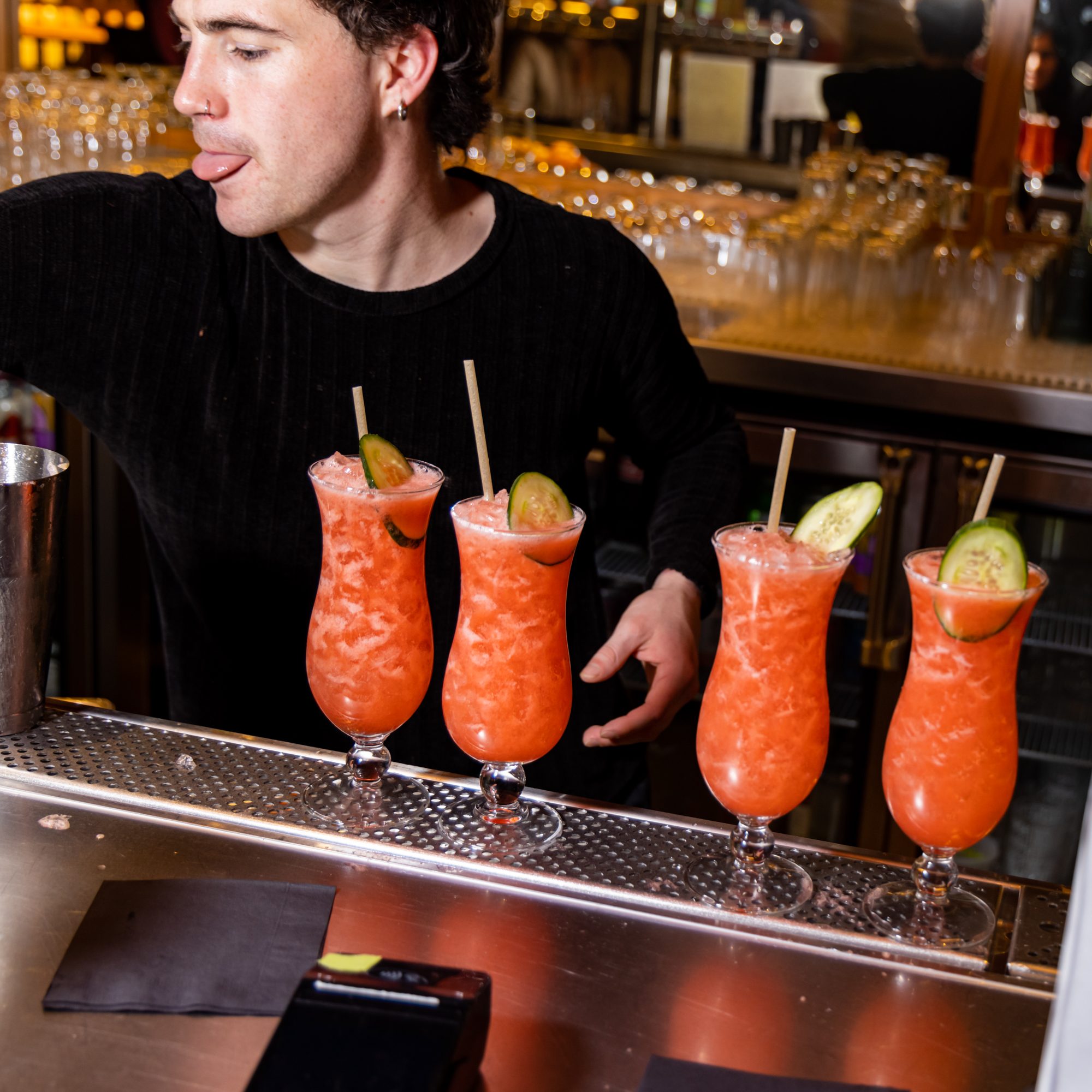 A bartender is preparing four bright orange cocktails, each garnished with a cucumber slice and a straw, on a stainless steel bar counter.