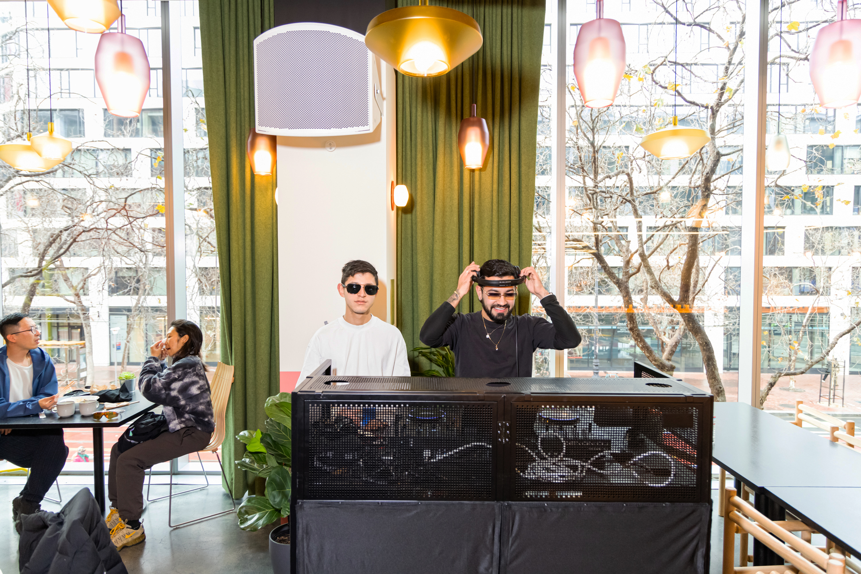 Two men stand behind DJ equipment in a cafe with large windows. One adjusts headphones, the other wears sunglasses. Diners are seated at tables nearby.