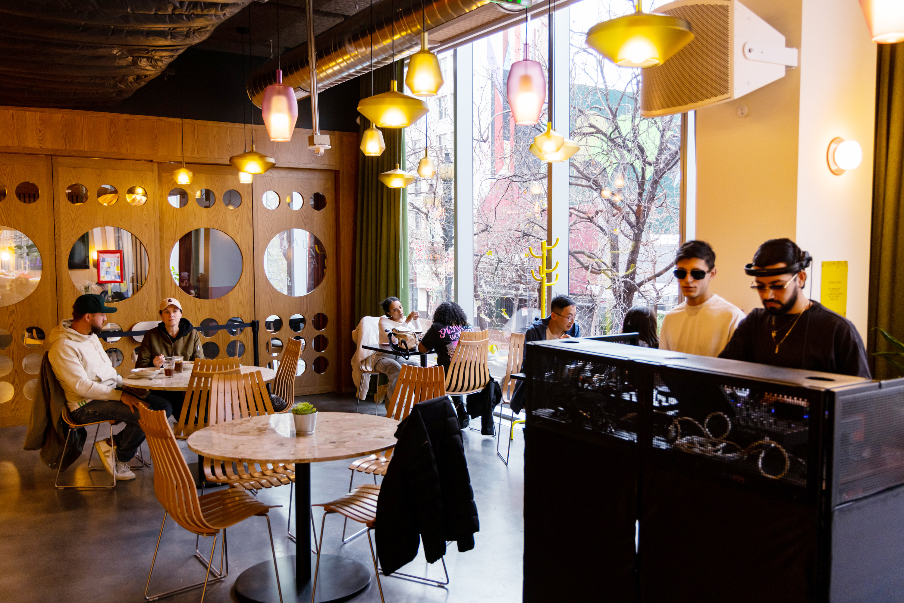 A cozy cafe with wooden decor features people sitting at tables, chatting and using laptops. Two individuals are at a DJ booth near large windows.