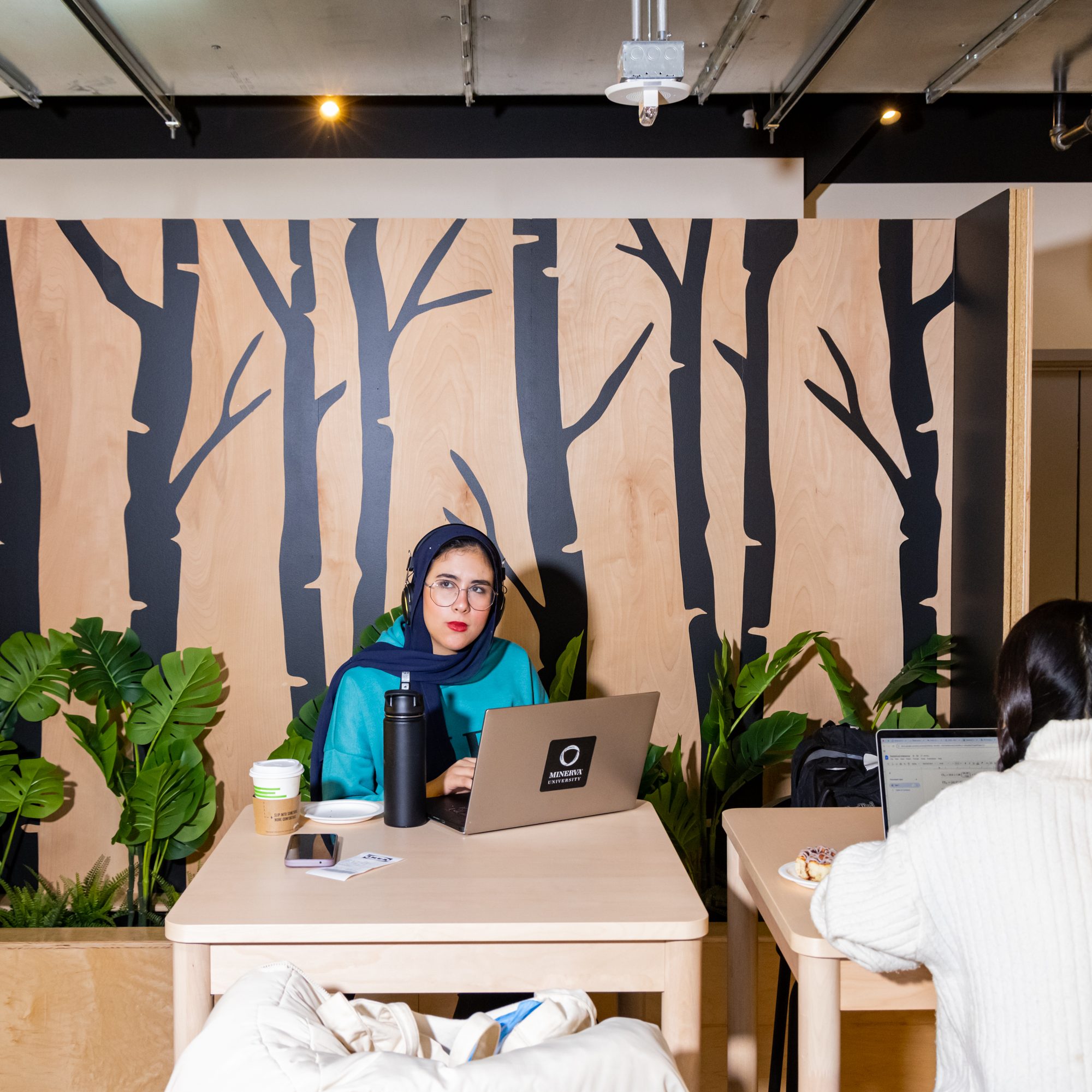 A person in a teal outfit sits at a table with a laptop, headphones, and a drink, set against a mural of black tree silhouettes. Another person works nearby.