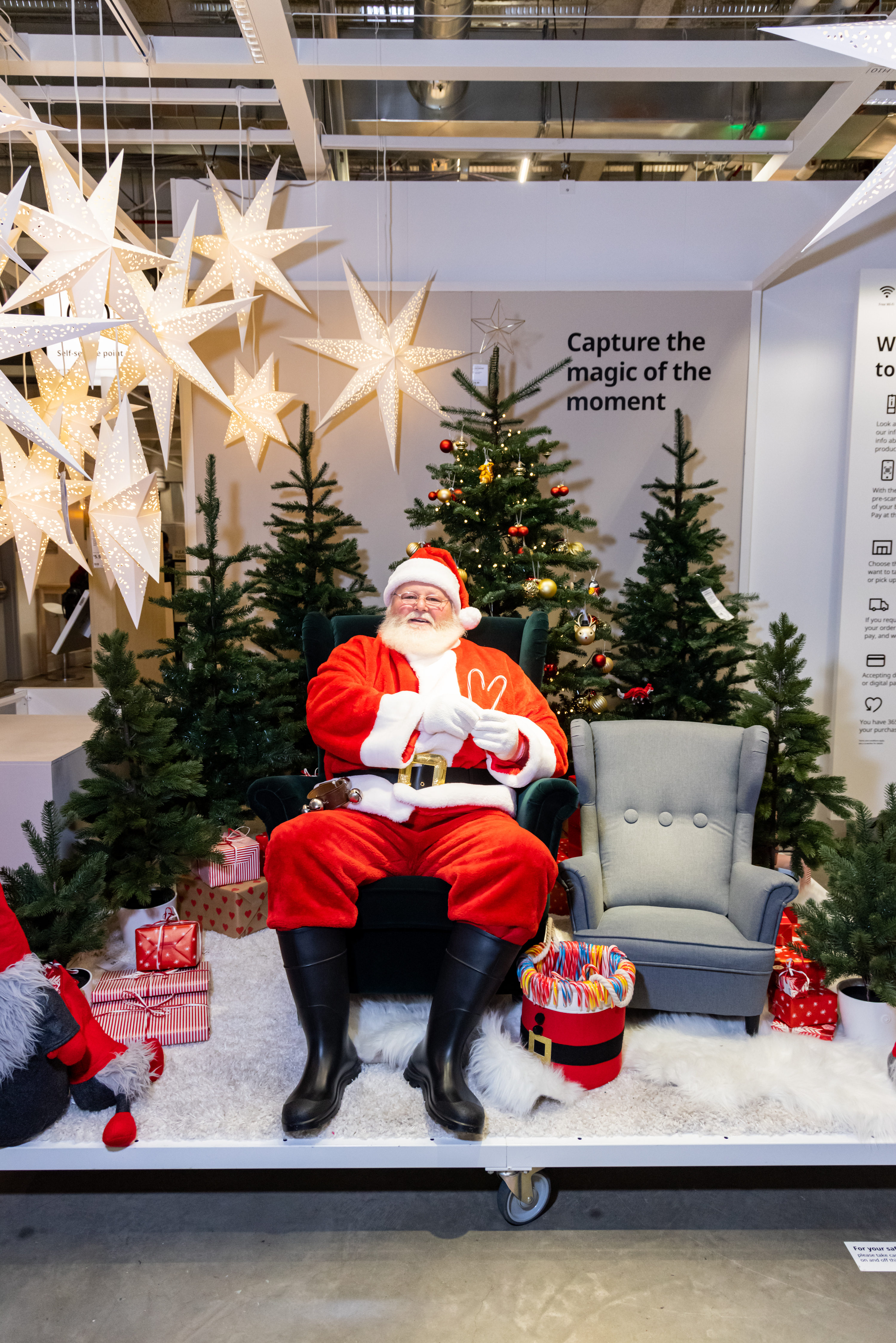 A person dressed as Santa sits on a green chair with a peace sign, surrounded by Christmas trees, star lights, and wrapped gifts on a white rug.