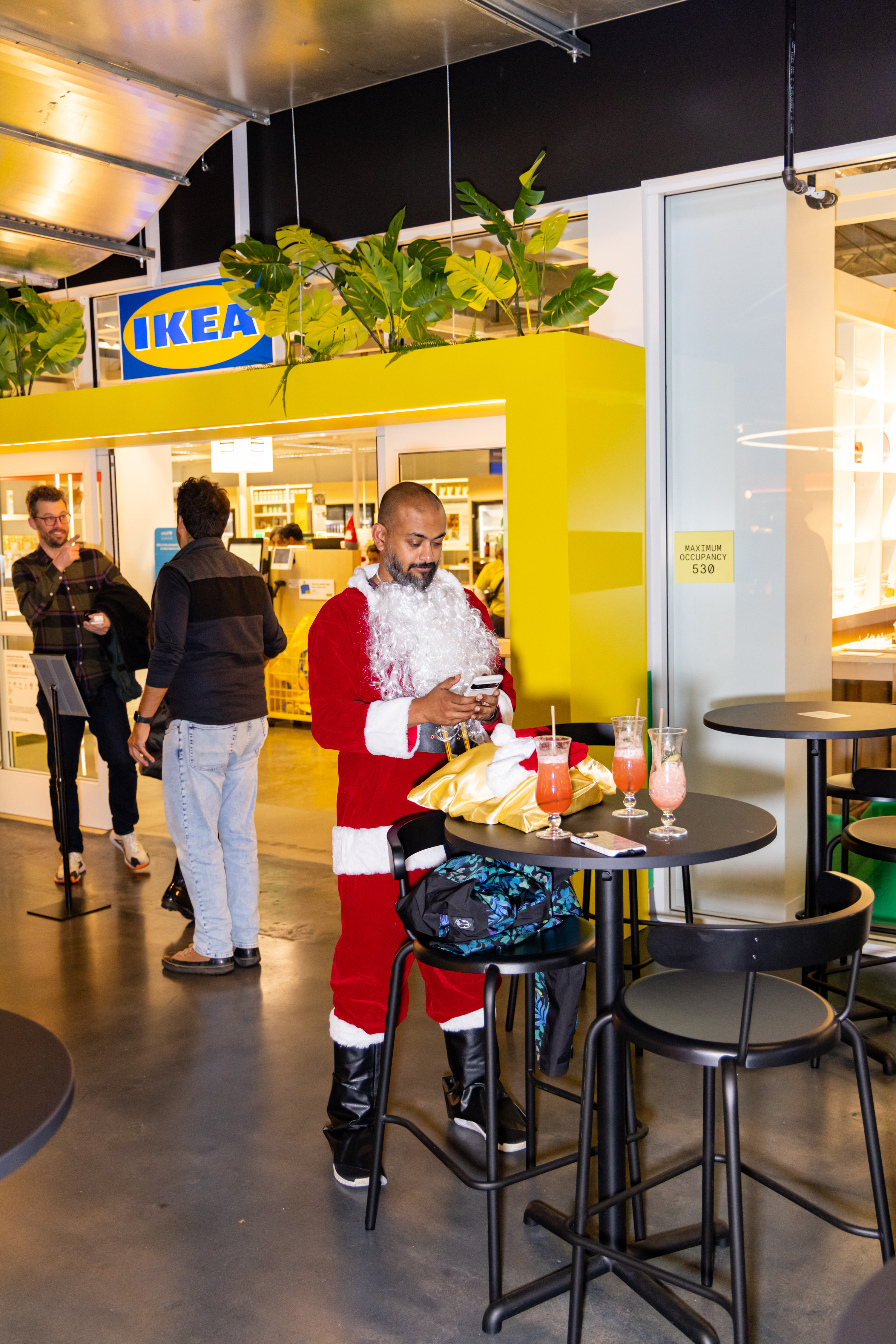 A man in a Santa suit is using a phone at a high table with drinks, inside an IKEA store. The store's yellow sign and green plants are visible above him.