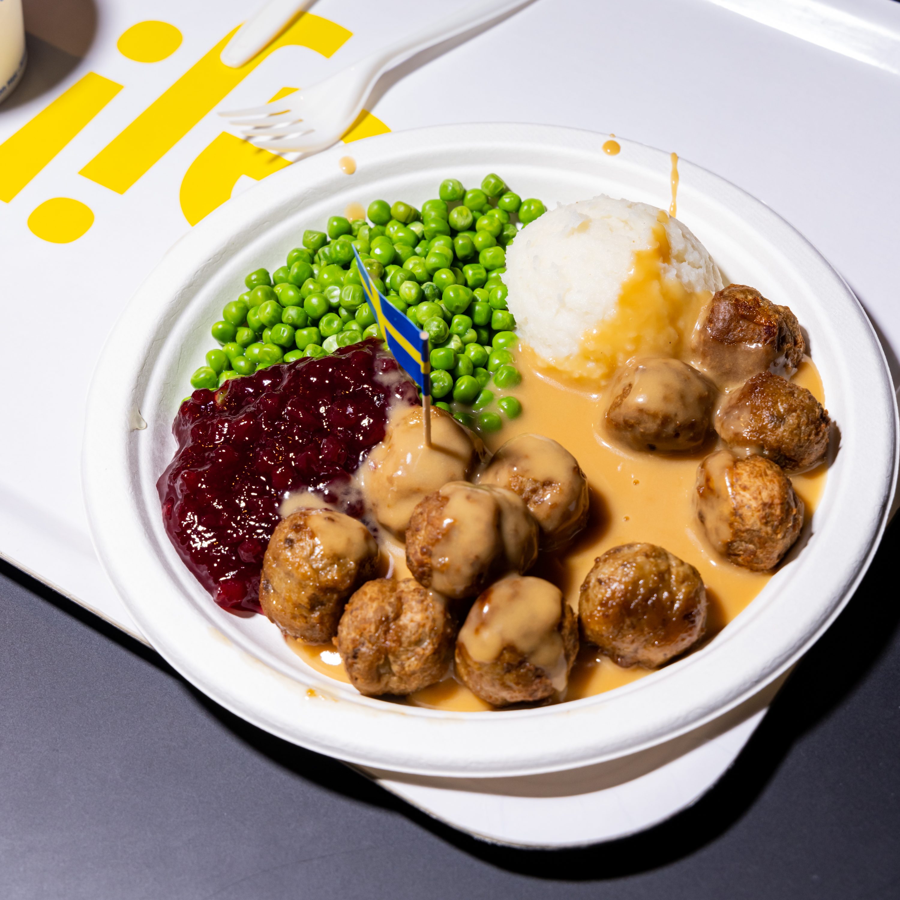 A plate of Swedish meatballs with gravy, mashed potatoes, green peas, and a dollop of lingonberry sauce, garnished with a small Swedish flag.