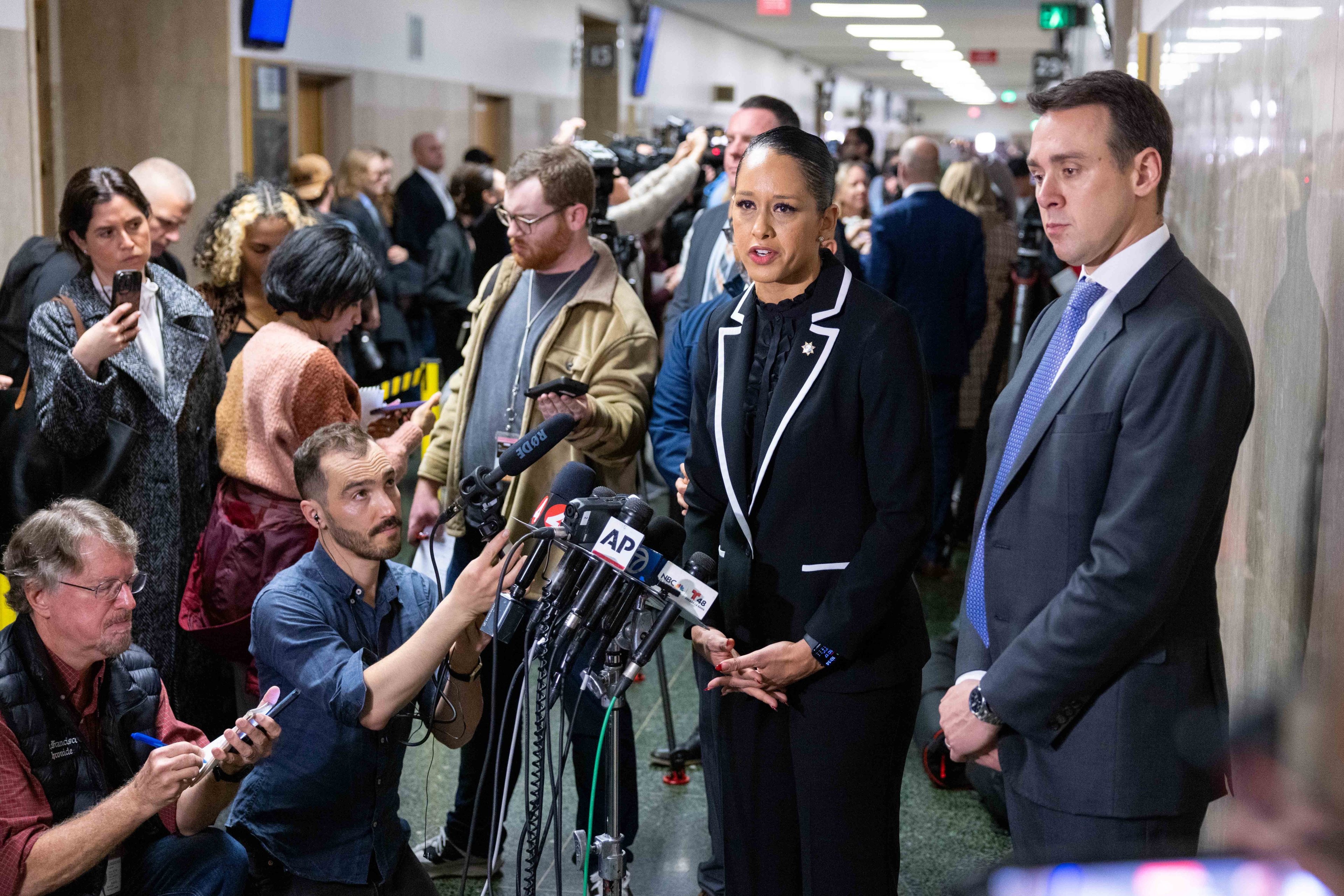 A woman in a suit inside a courthouse takes questions from reporters pointing microphones.