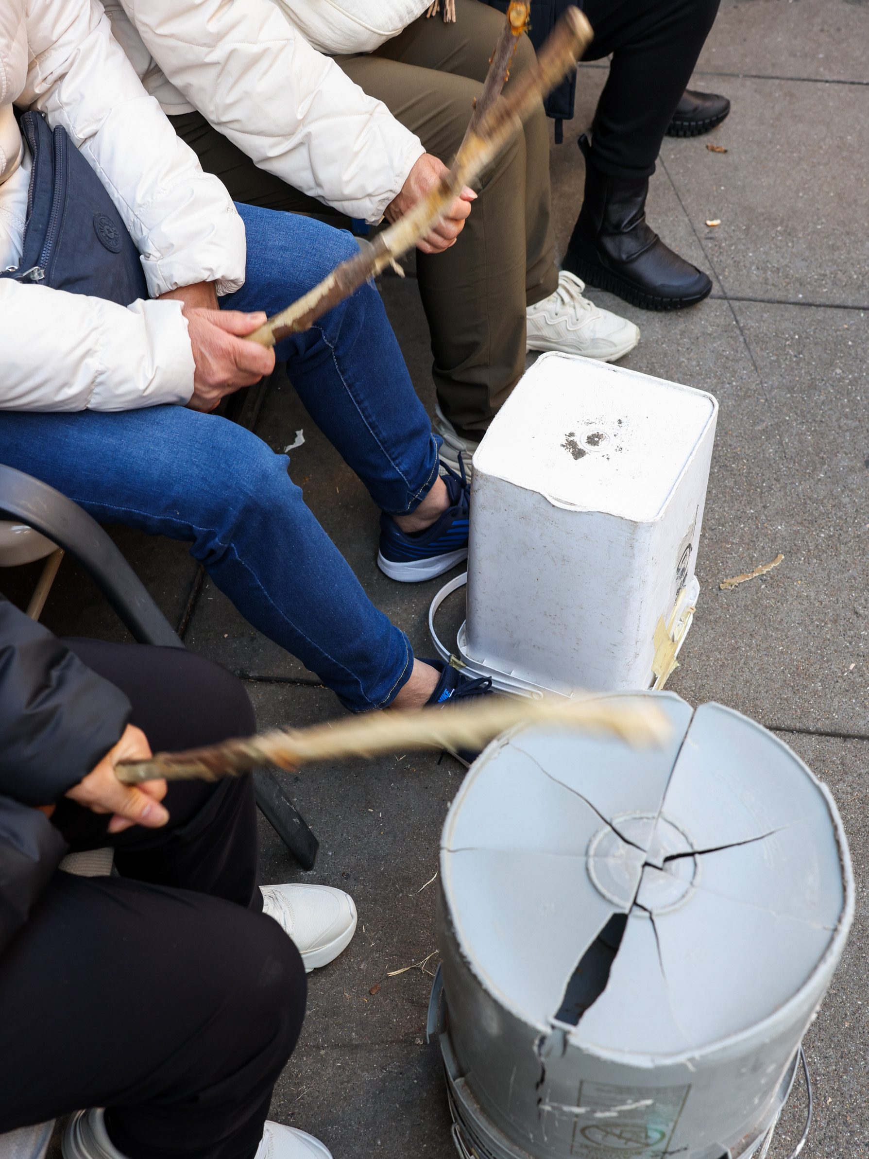 Two people in white jackets are seated and playing drums with wooden sticks. The drums are makeshift from plastic buckets, placed on the ground.