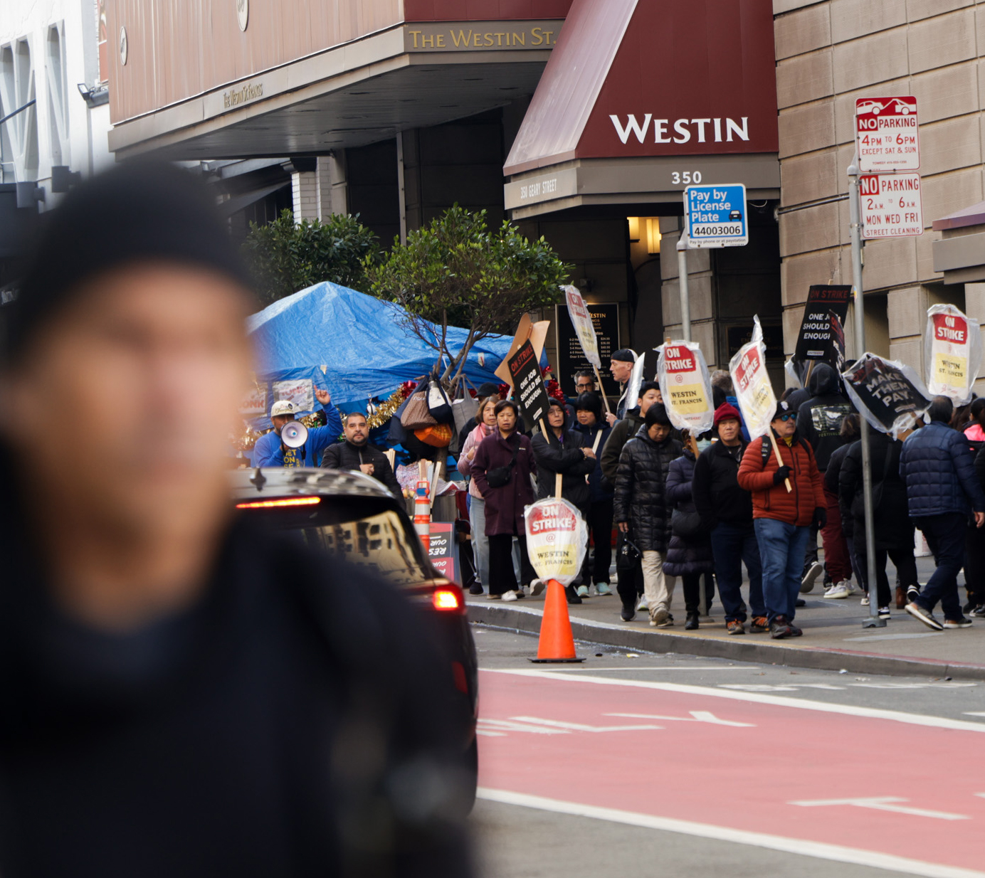 A group of people is picketing outside a building with &quot;Westin&quot; signage, holding strike signs with messages like &quot;On Strike&quot; and &quot;Make Them Pay.&quot;